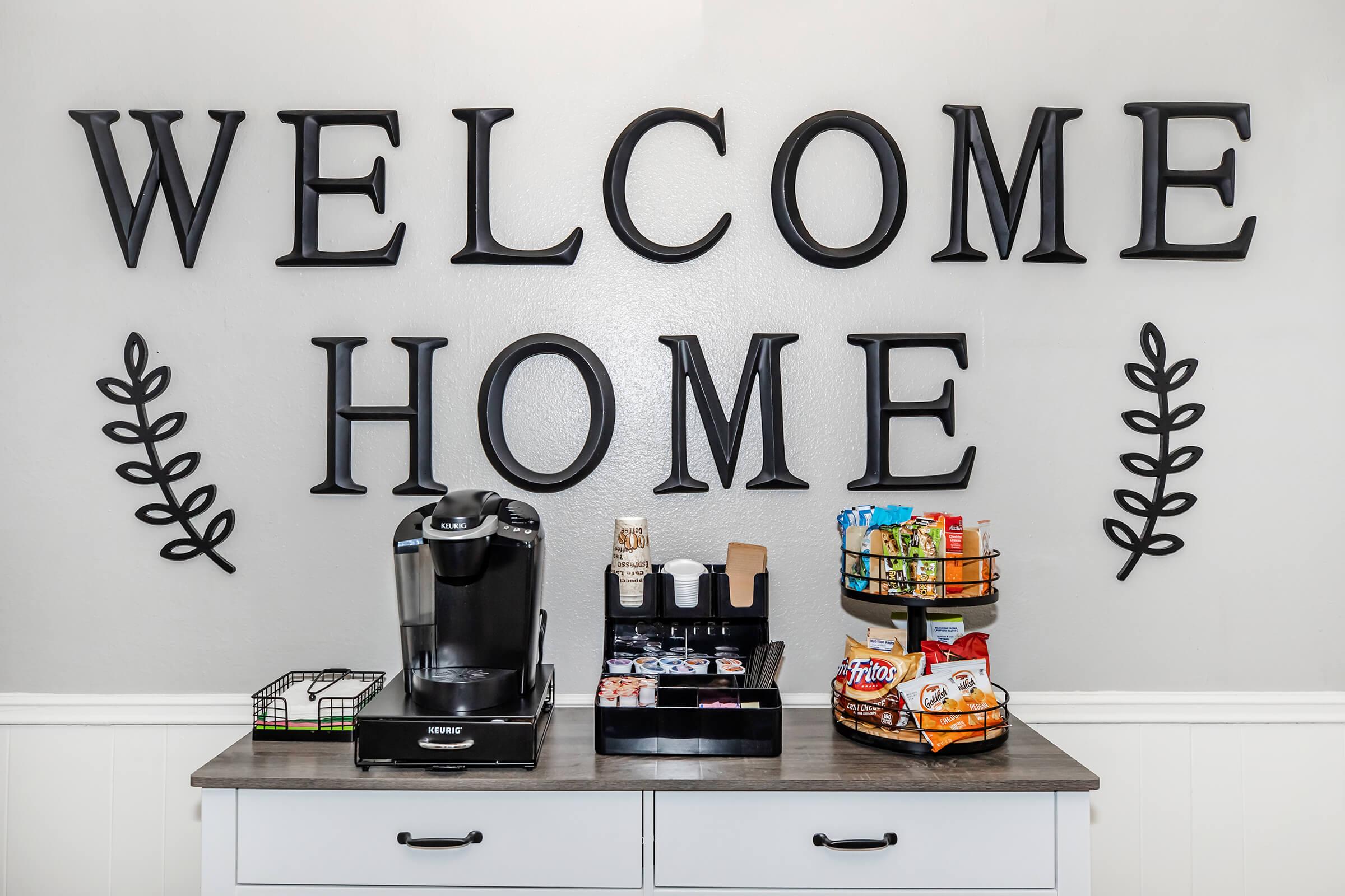 Coffee station with a Keurig machine, a selection of coffee pods, and various snacks displayed on a tiered rack. The wall features the words "WELCOME HOME" in large letters, surrounded by decorative leaf motifs. The overall aesthetic is warm and inviting.