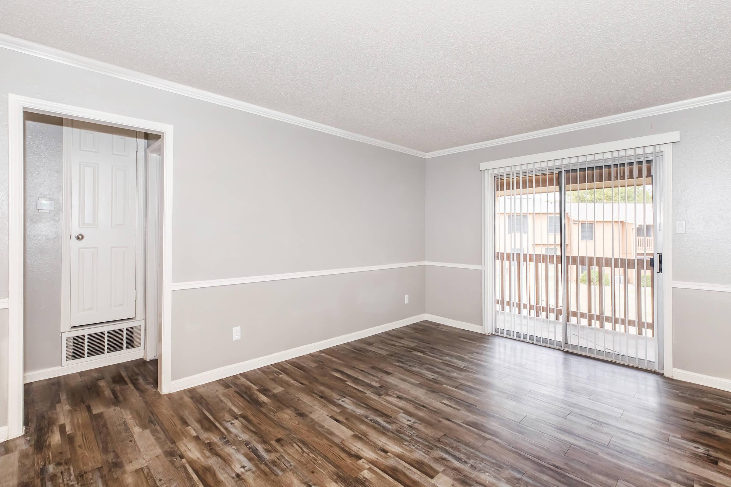 Interior view of a spacious living room featuring gray walls, wooden flooring, and a sliding glass door leading to a balcony. A white door on the left leads to another room, and there is baseboard heating. The overall atmosphere is bright and airy, ideal for a cozy living space.