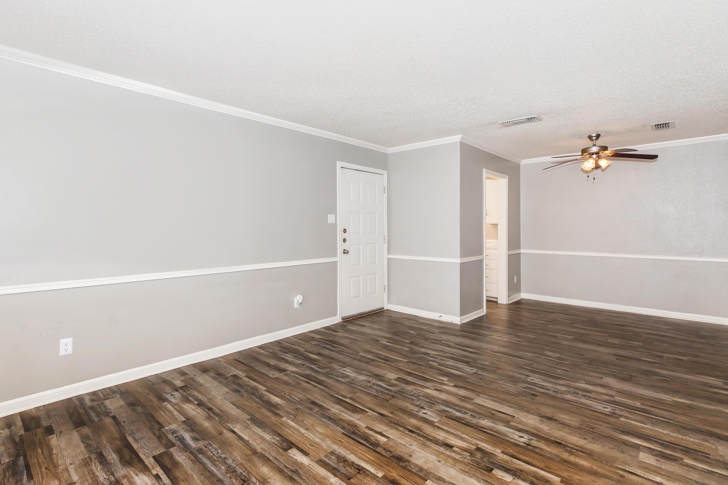 Spacious living room with light gray walls and a white trim. Features a ceiling fan, a front door, and a view into a kitchen area. The floor is covered in dark wood planks, creating a modern and inviting atmosphere. Bright lighting enhances the open space.