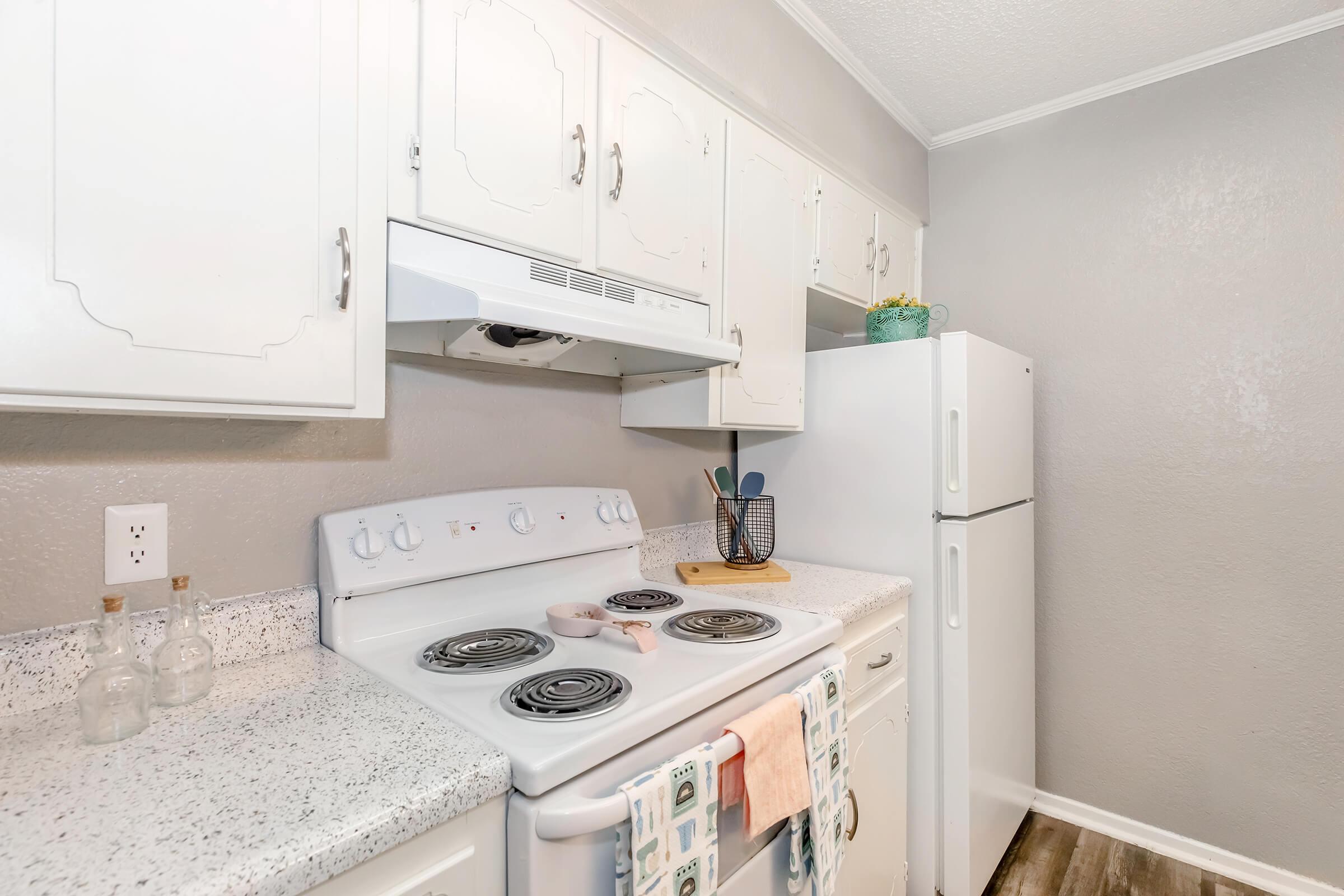 A modern kitchen featuring white cabinetry, a stovetop with four burners, and an overhead exhaust fan. The countertops are speckled gray and include a cutting board and utensils. A refrigerator is present, along with decorative elements like a jar and plants on the shelves. The walls are painted a light gray.