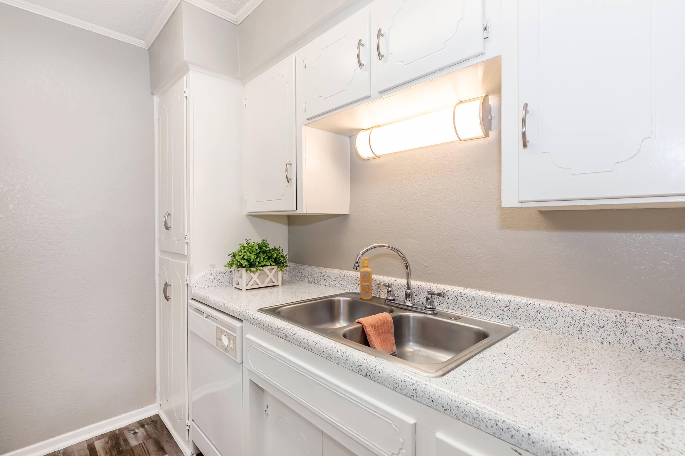 A clean kitchen with white cabinets, a sink with a faucet, and a small potted plant on the counter. There's a dish towel resting on the edge of the sink, and the countertops are speckled. A light fixture above the sink provides illumination, creating a bright and inviting atmosphere.
