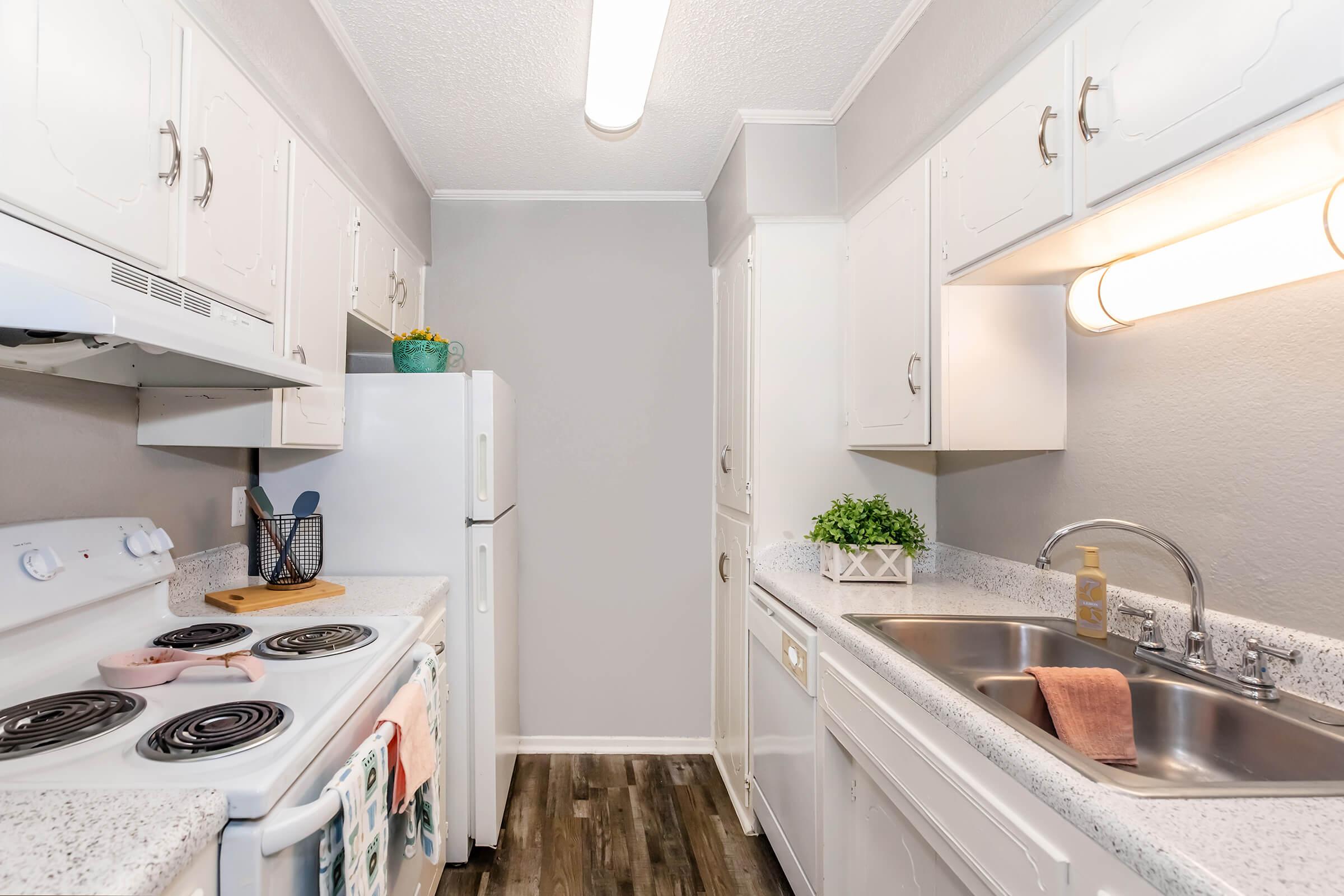 A modern kitchen featuring white cabinets, a stove with a curved hood, a refrigerator, and a double sink. The countertop is light gray with a small plant and kitchen utensils visible. Soft lighting illuminates the space, which has gray walls and a wooden floor, creating a clean and inviting atmosphere.