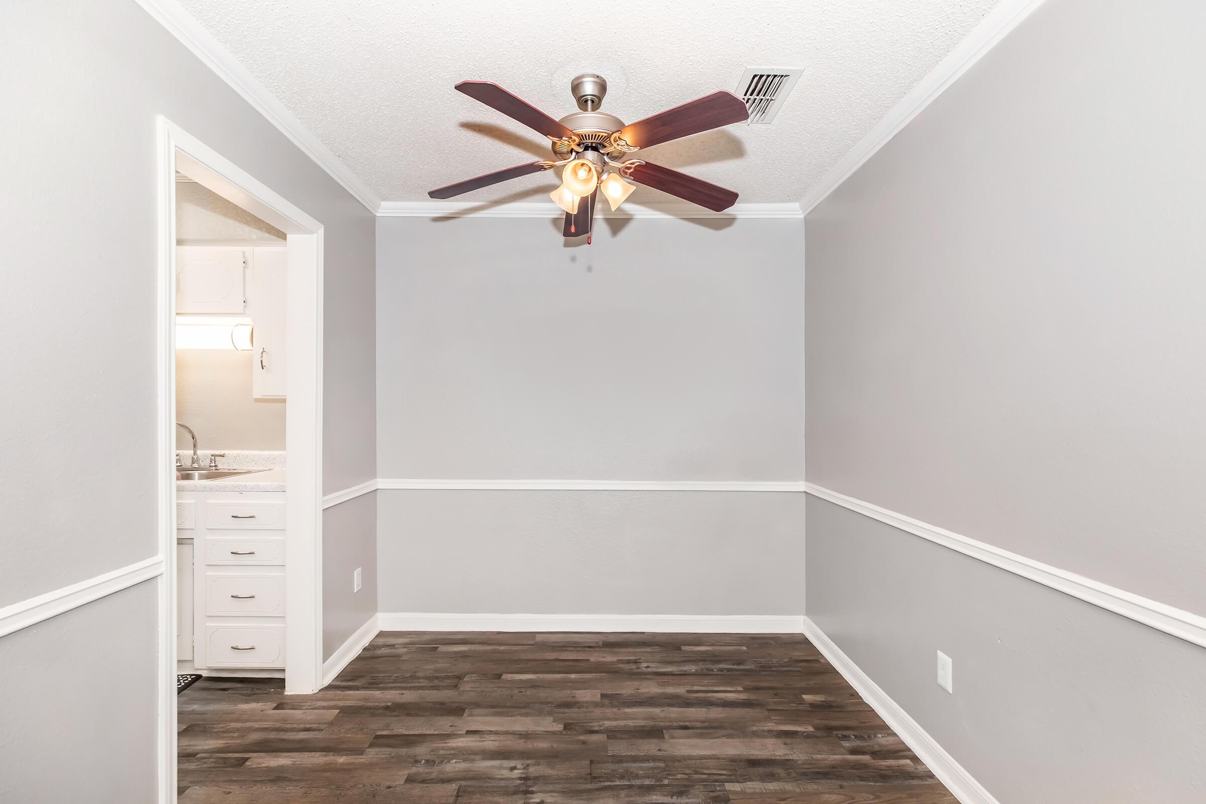 A blank room featuring gray walls and a ceiling fan with four lights. The floor is made of dark wood, and there is a doorway leading to another area on the left side. The interior has a clean and modern appearance, with a simple white trim along the baseboards.