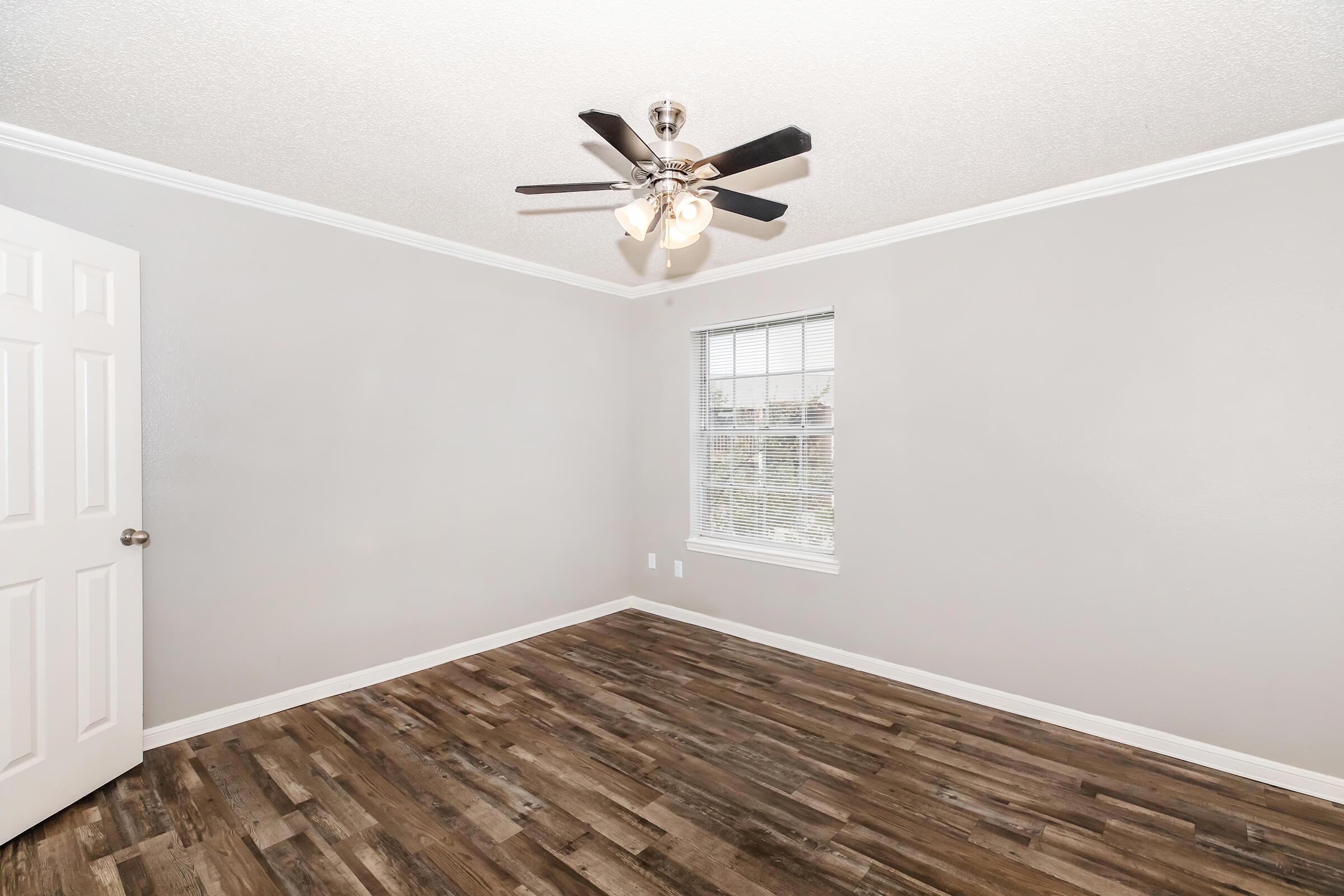A well-lit empty room with light gray walls, a ceiling fan with dark blades, and a window with blinds. The floor features wooden planks, and there is a white door visible on the left side of the image.