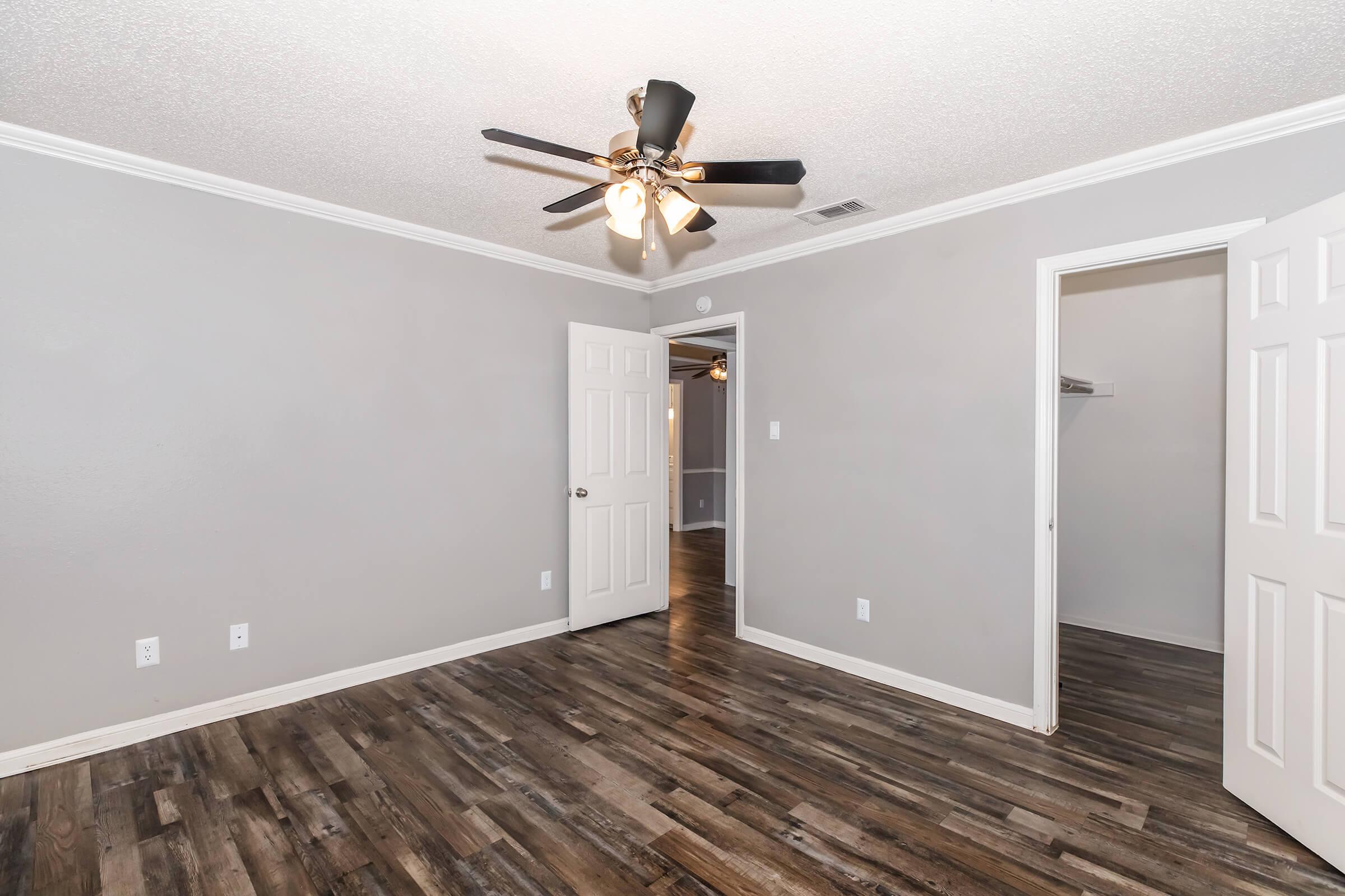 A spacious, neutral-toned room with a ceiling fan. The walls are painted gray, and the floor features dark wood laminate. A white door leads to an adjoining room or closet, while another door opens to a hallway. The room is well-lit and empty, ready for personal touches or furnishings.