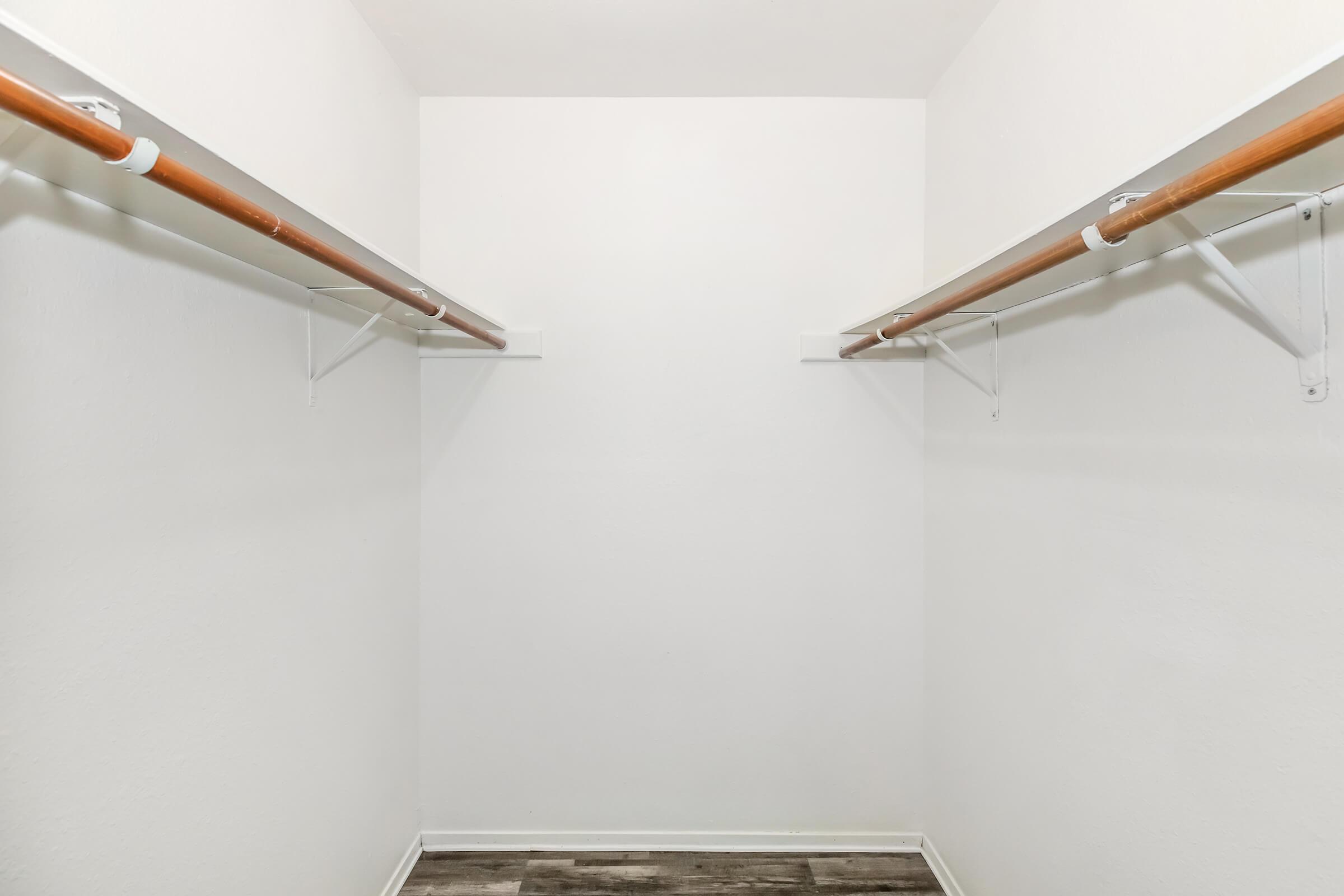 Empty closet interior with two hanging rods and white walls. The floor features a neutral-colored laminate. The space is well-lit and uncluttered, providing a minimalist view of the storage area.