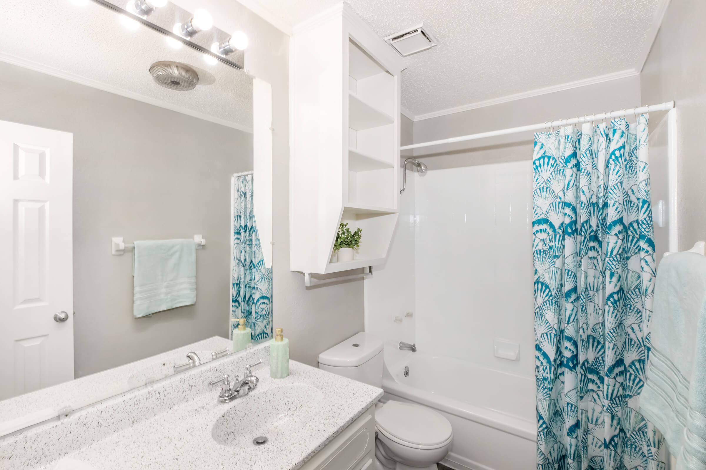 A modern bathroom featuring a clean design with light gray walls, a white bathtub with a shower curtain adorned with a blue aquatic pattern, a white vanity with a speckled countertop, and a large mirror. There are soft blue towels and a shelf storing decorative items, creating a fresh and inviting atmosphere.