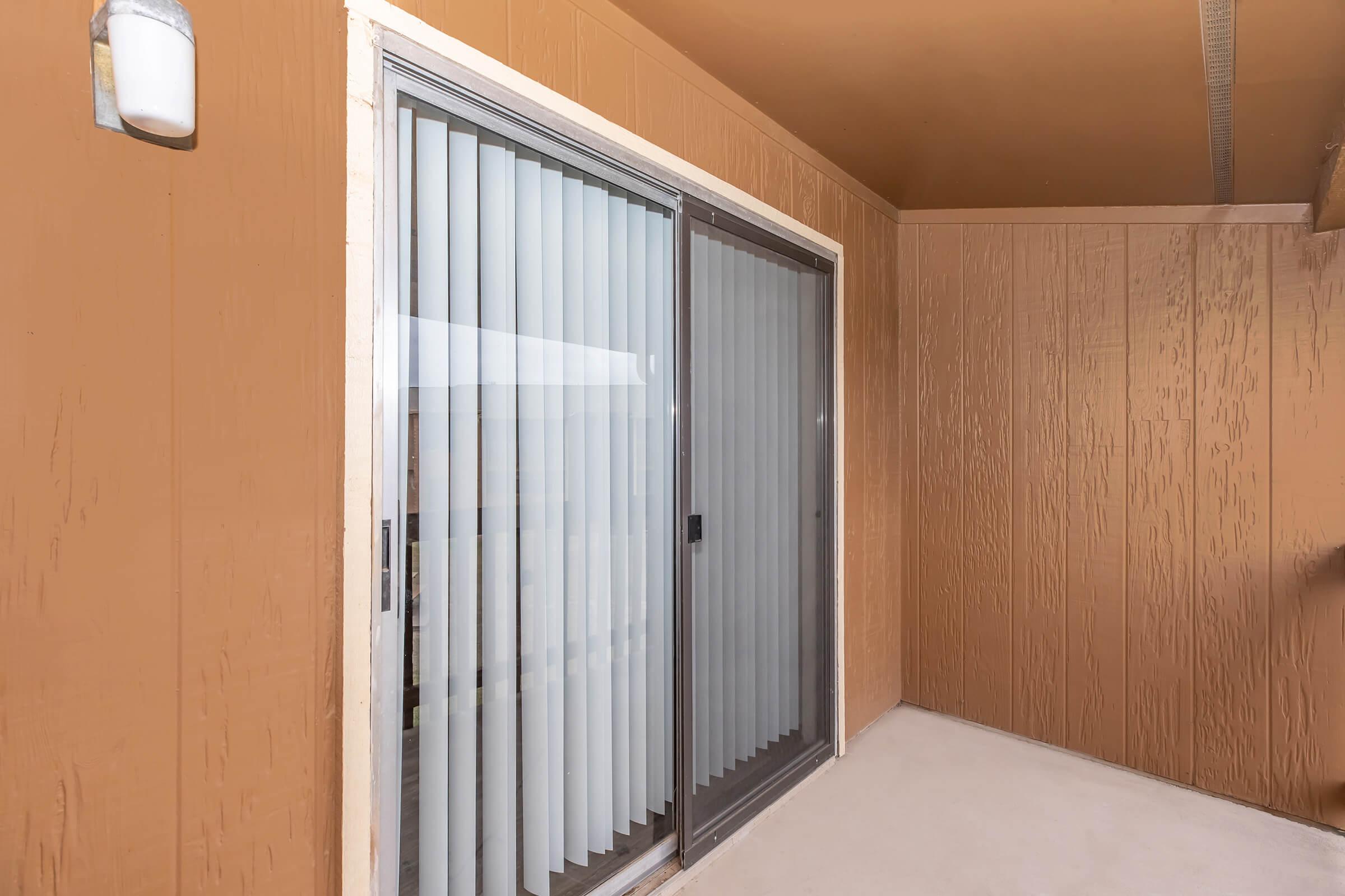 Sliding glass doors with vertical blinds open to a balcony area. The walls are painted a warm brown, and there is a light fixture on the left. The floor is a light-colored surface, and the overall ambiance is bright and inviting.