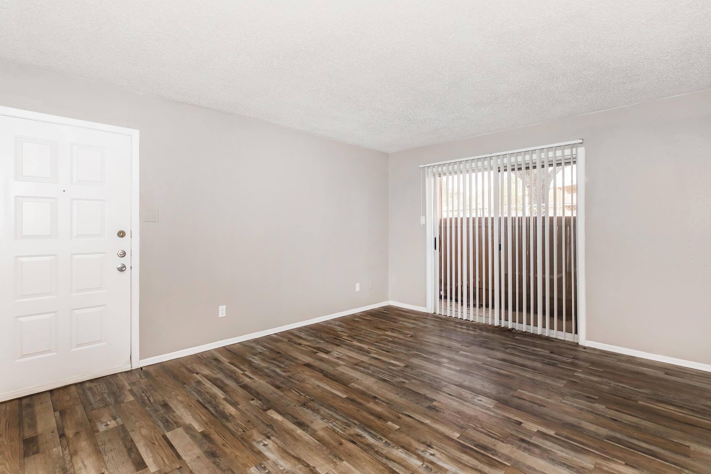 A spacious, empty living room featuring light-colored walls and a modern, wood-like laminate floor. The left side shows a white front door, while the right side has a sliding glass door with vertical blinds, leading to an outdoor area. Natural light fills the room, creating a bright and airy atmosphere.