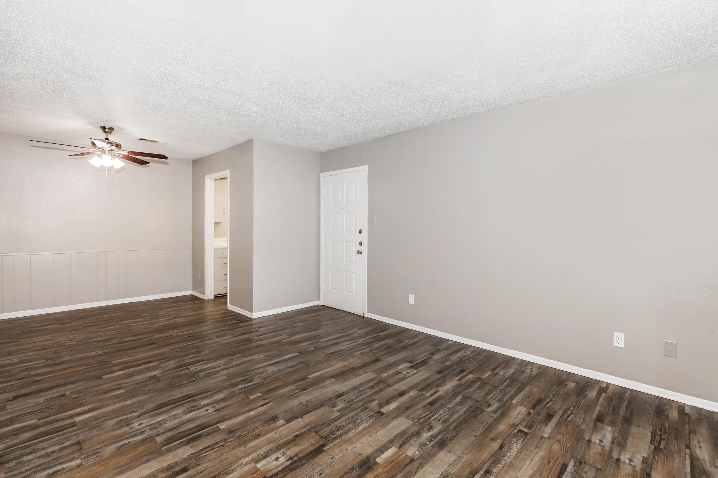 A spacious, empty living room featuring light gray walls and wooden flooring. On the left, there is a doorway leading to a kitchen space, and in the center, a ceiling fan is visible. The entrance door is prominently displayed on the right side of the image. The overall ambiance is bright and inviting.