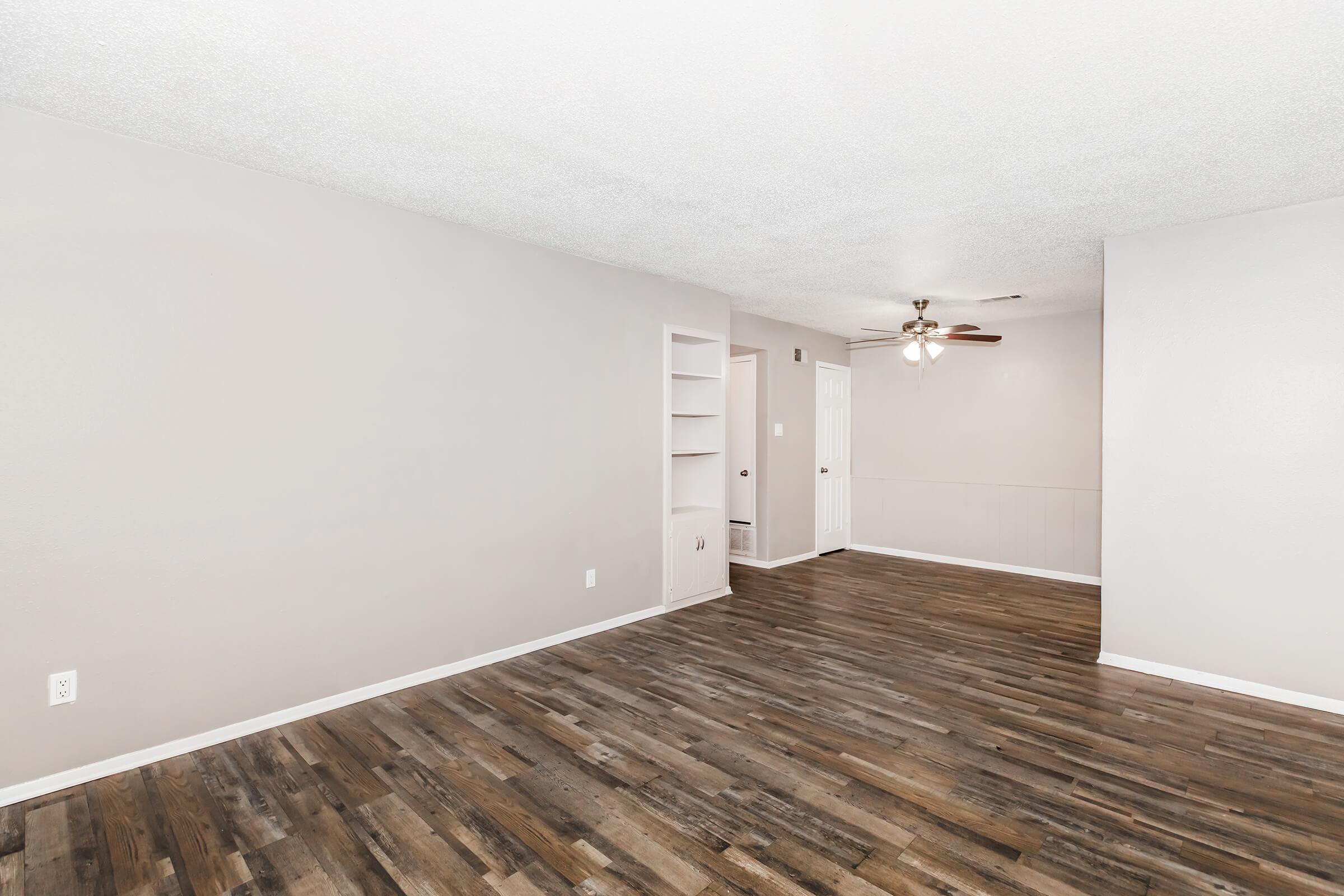 An empty, well-lit room with light gray walls and dark wooden flooring. There is a small built-in shelf on the left wall and a ceiling fan above. A door leads to another room on the right, and the space is free of furniture, creating an open and airy atmosphere.