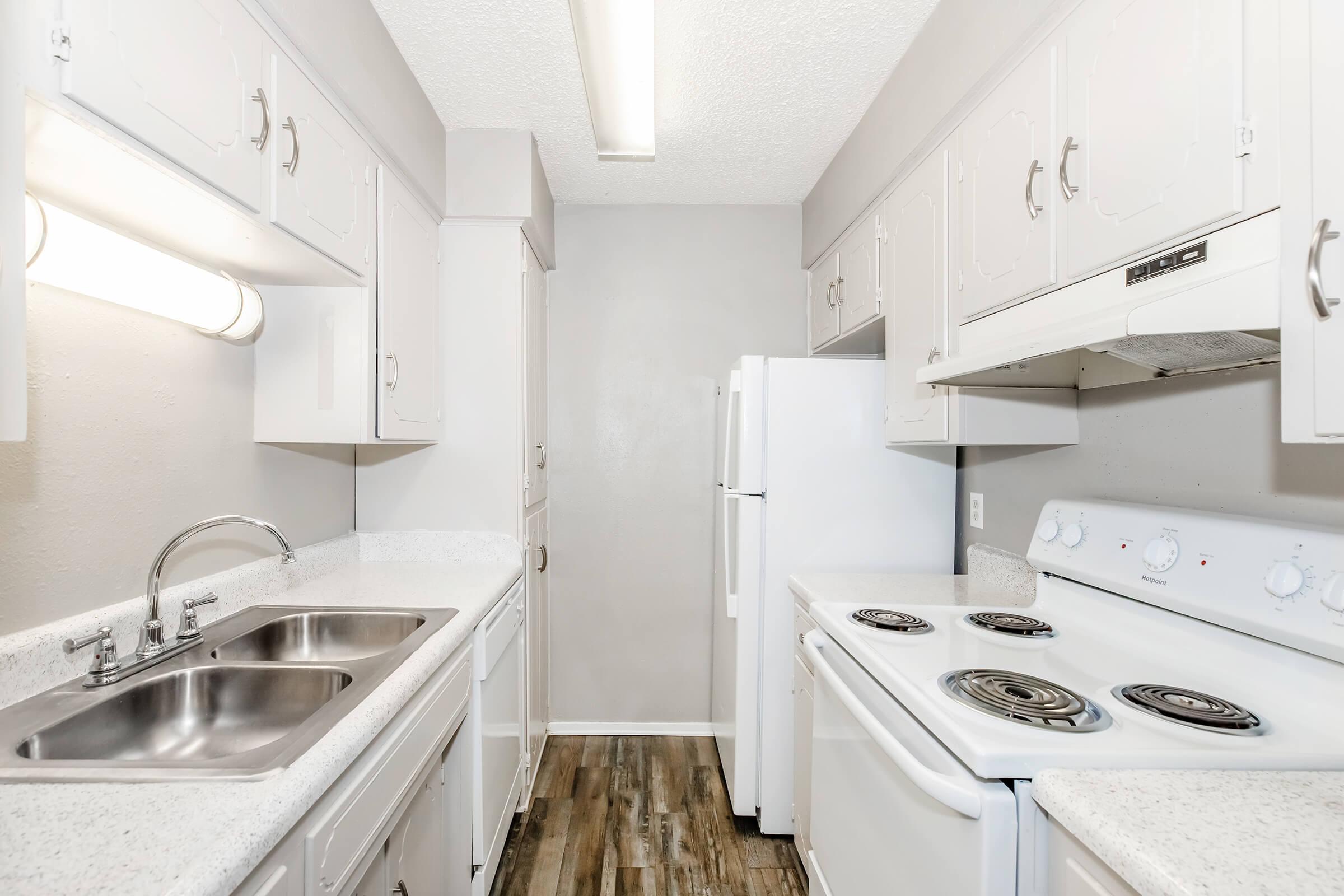 A clean, modern kitchen featuring white cabinetry, a double sink, a refrigerator, a stove with an oven, and laminate flooring. The walls are painted in a light gray color, and overhead lighting brightens the space.