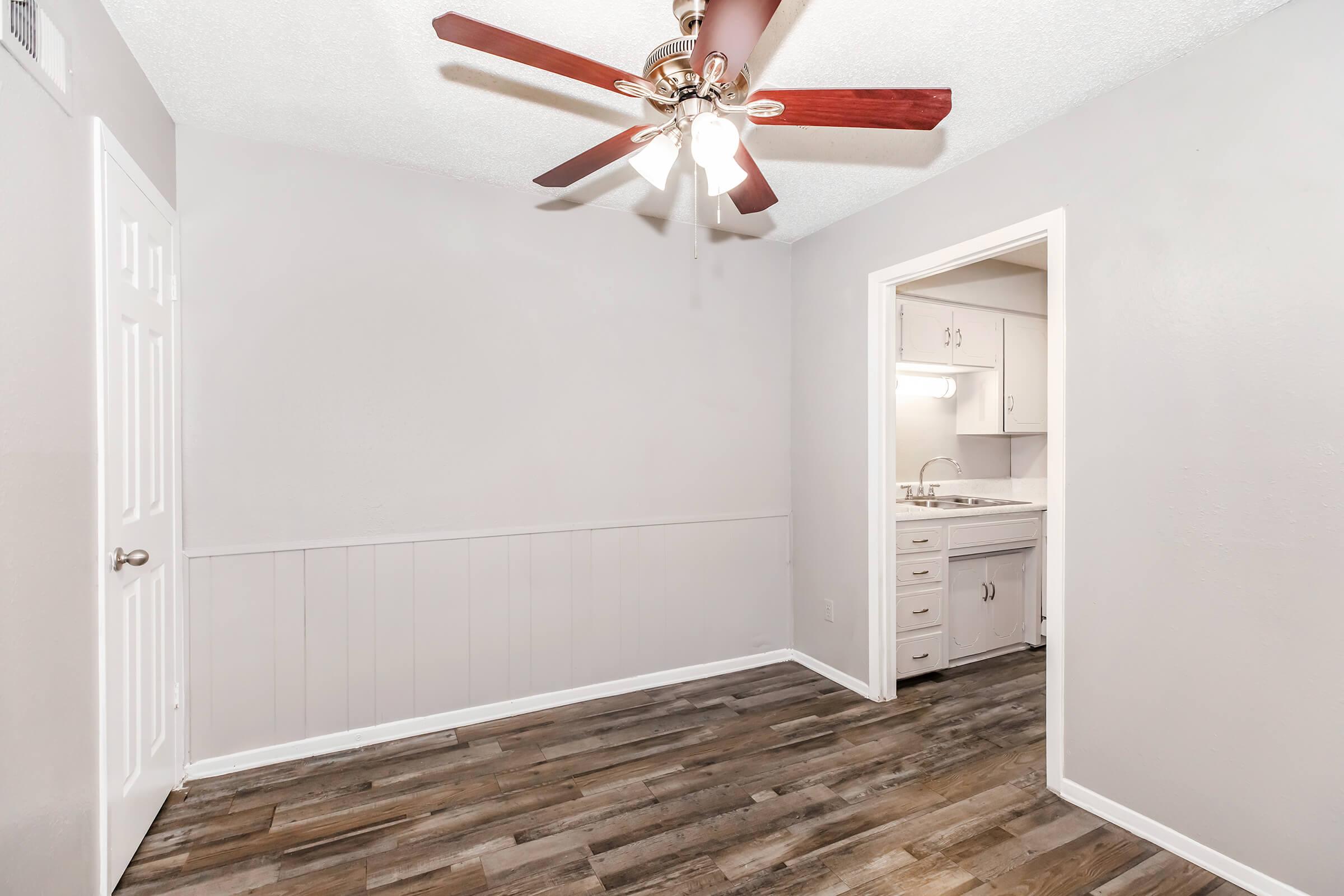 A small, empty room with light gray walls and wooden flooring. A ceiling fan with wooden blades is overhead. On one side, there is a doorway leading to a bathroom area with white cabinetry and a mirror, partially visible. The space is well-lit and clean, suitable for various uses.