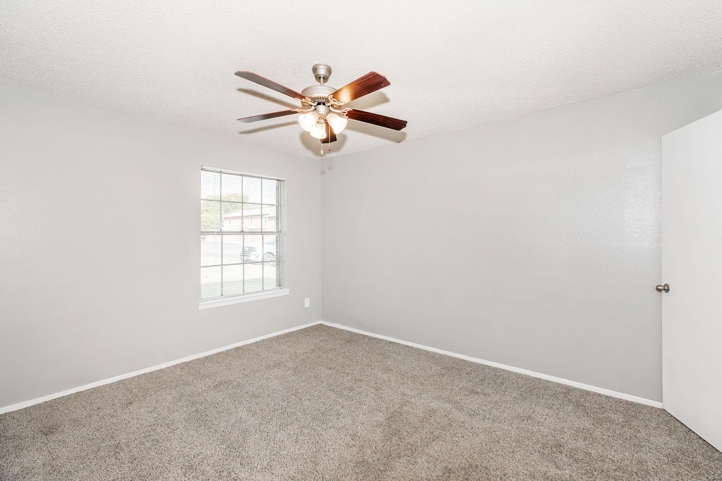 A spacious, empty room with light gray walls and plush carpet. A ceiling fan with wooden blades is visible, and there is a window allowing natural light to enter. A white door is shown on the right, adding to the clean and simple aesthetic of the space.