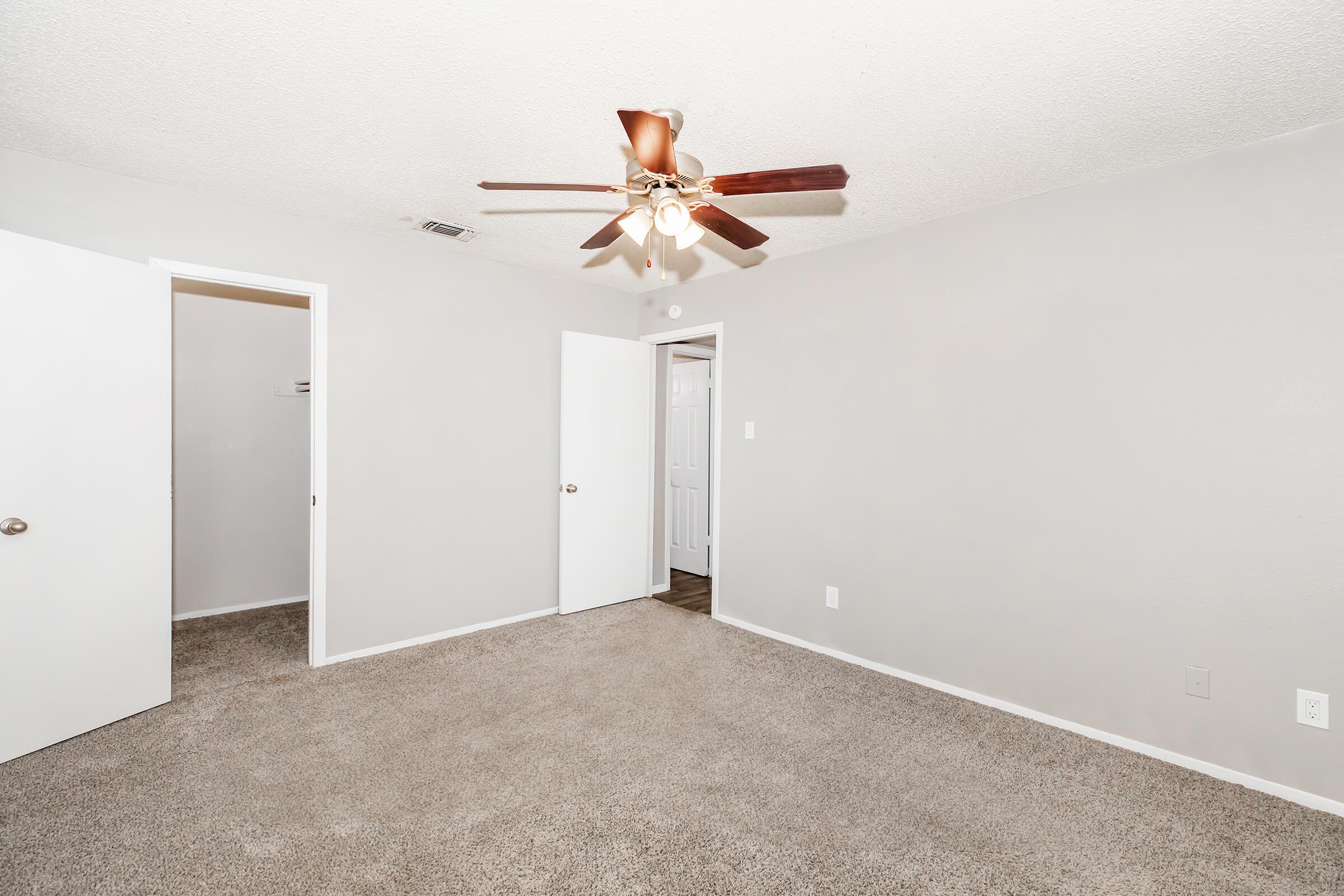 A spacious, empty bedroom with light gray walls and plush carpet. It features a ceiling fan with wooden blades and two doors leading to closets or other rooms. The room is well-lit with natural light, creating a clean and inviting atmosphere.