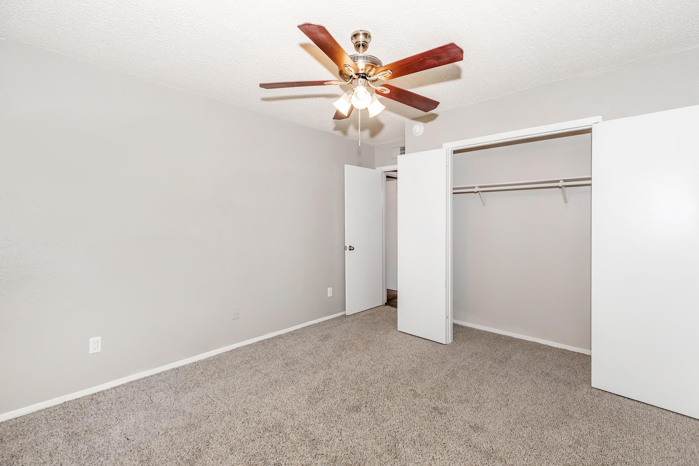 A small, well-lit bedroom featuring light gray walls and carpet. It has a ceiling fan with wooden blades. One side of the room has a white closet with double doors, and an additional door leads to another area, possibly a hallway. The space appears clean and inviting.
