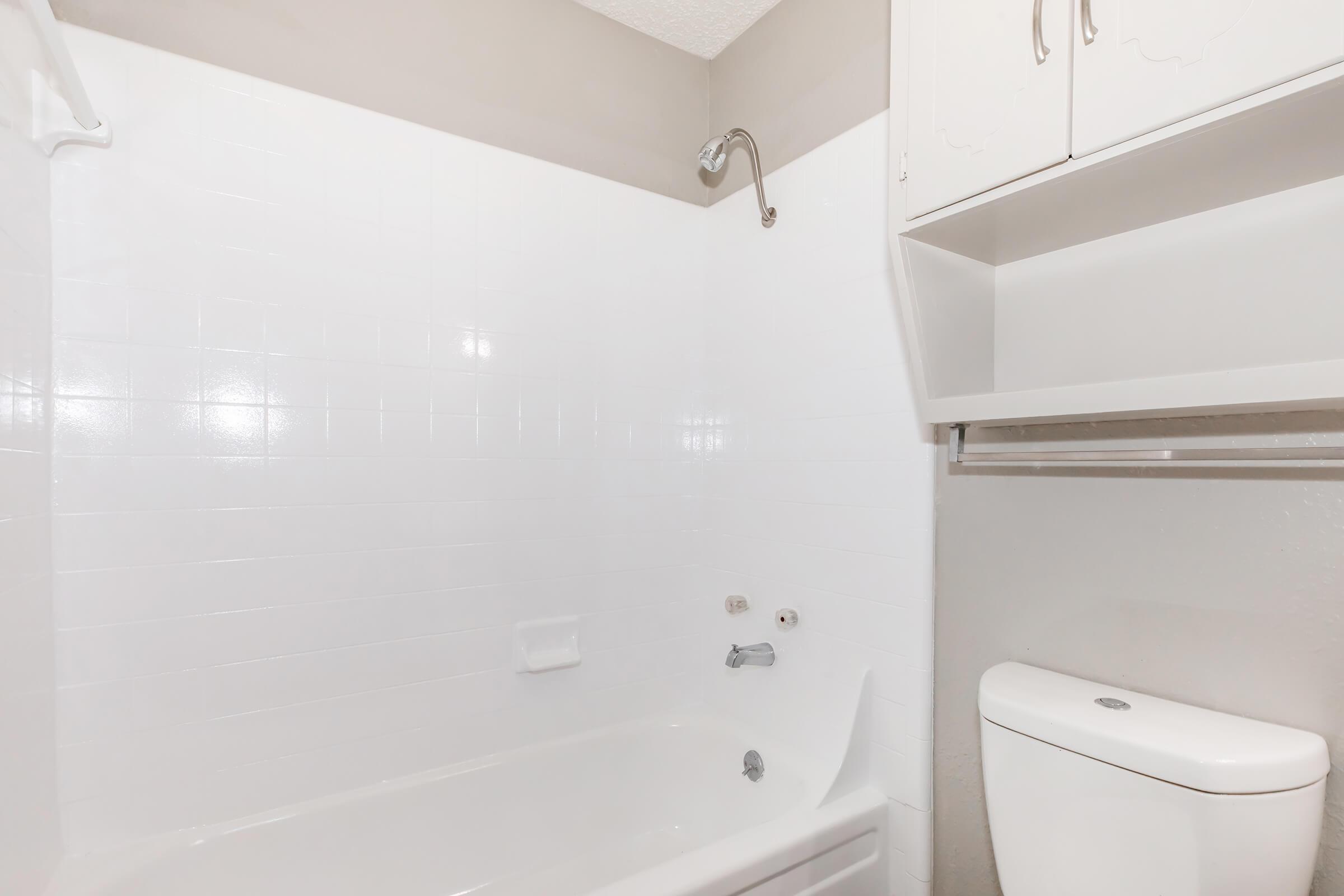 A clean and simple bathroom featuring a white bathtub with a chrome faucet, a wall-mounted showerhead, and a toilet. The walls are tiled in white, creating a bright and open space. Above, there is a cabinet for storage, complementing the minimalist design.