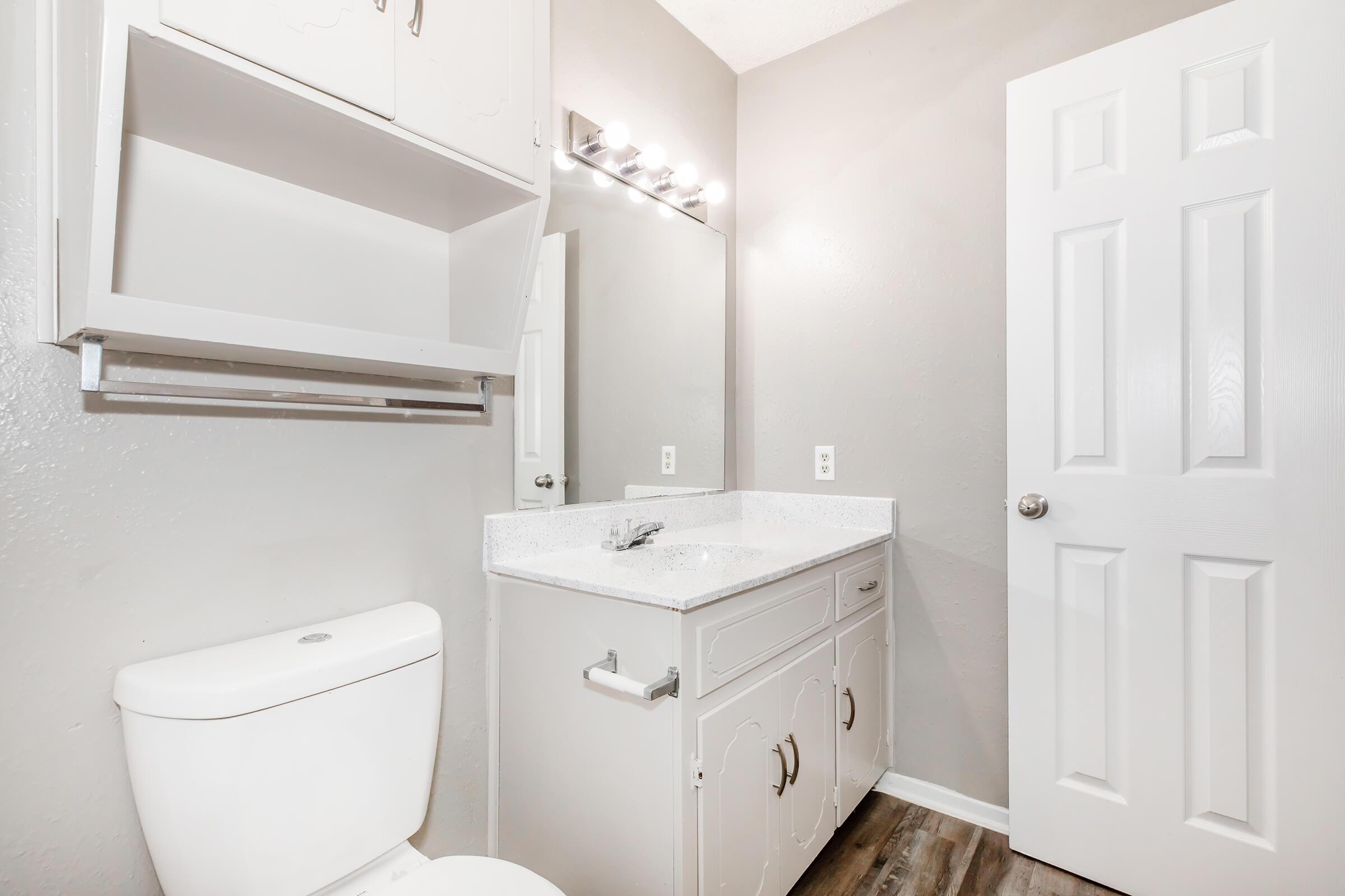 A modern bathroom featuring a white toilet and a vanity with a granite countertop. A large mirror is illuminated by overhead lights, and the walls are painted a soft gray. A cabinet above the sink provides storage, and a door leads to another room. The flooring appears to be a wood-like laminate.