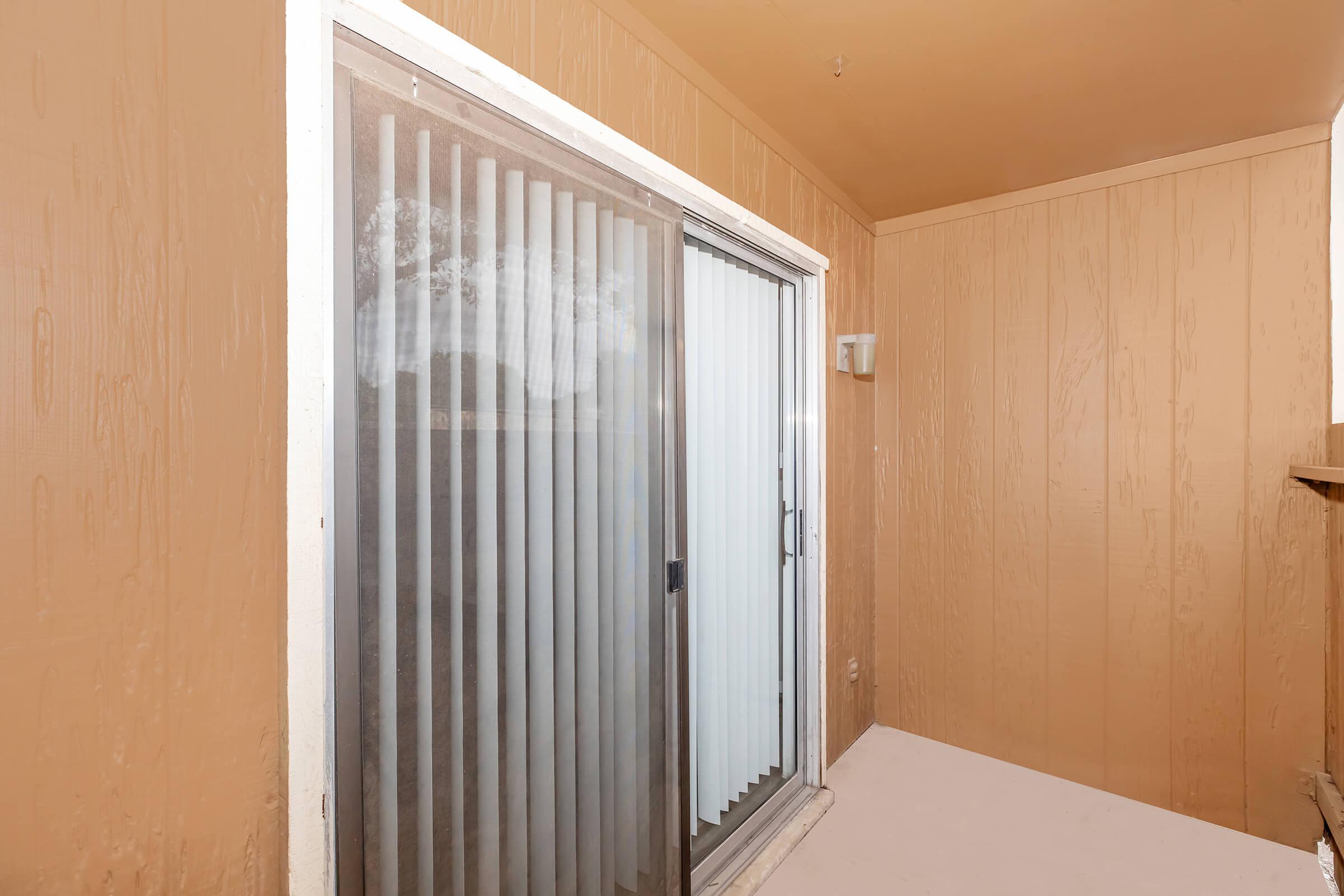A small balcony or patio area with beige walls, featuring a sliding glass door with vertical blinds. The floor is plain and the space appears well-lit. A light fixture is visible on the wall, adding to the simple, functional aesthetic of the area.