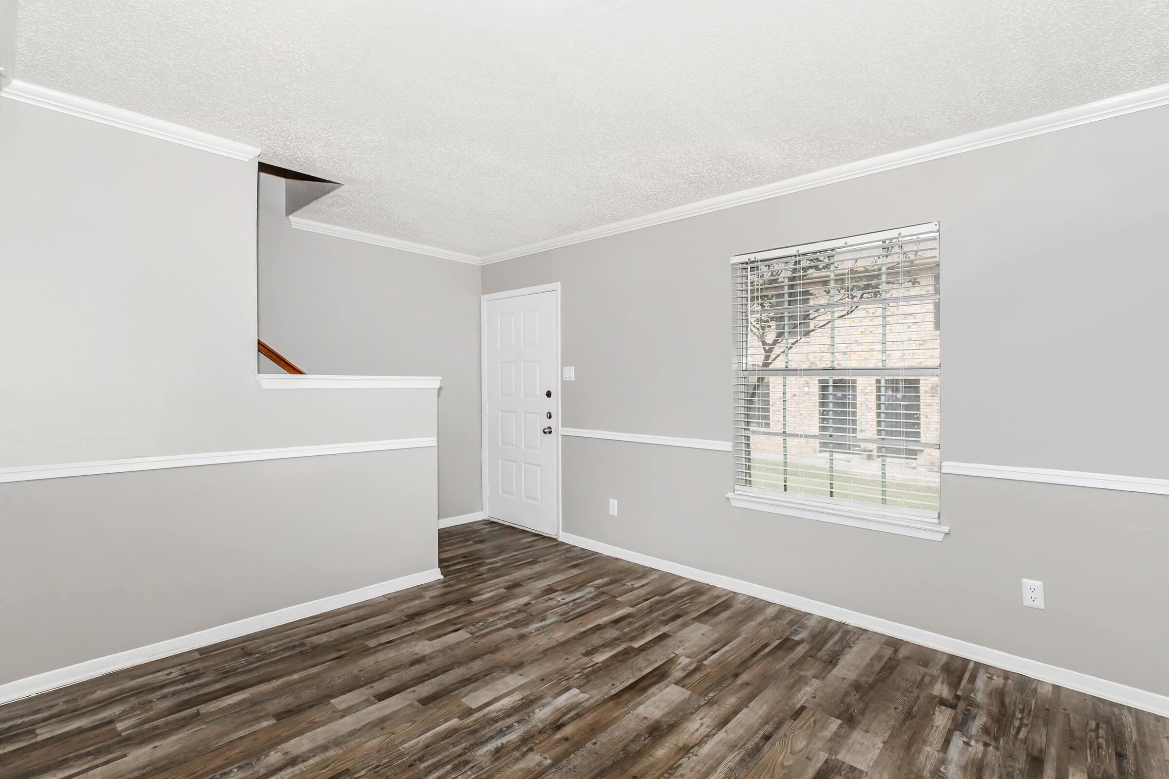 Interior view of a living space featuring light gray walls, a white baseboard, and a wood-like floor. A staircase is visible in the background, and there's a door leading outside. A window is present, allowing natural light to enter the room.