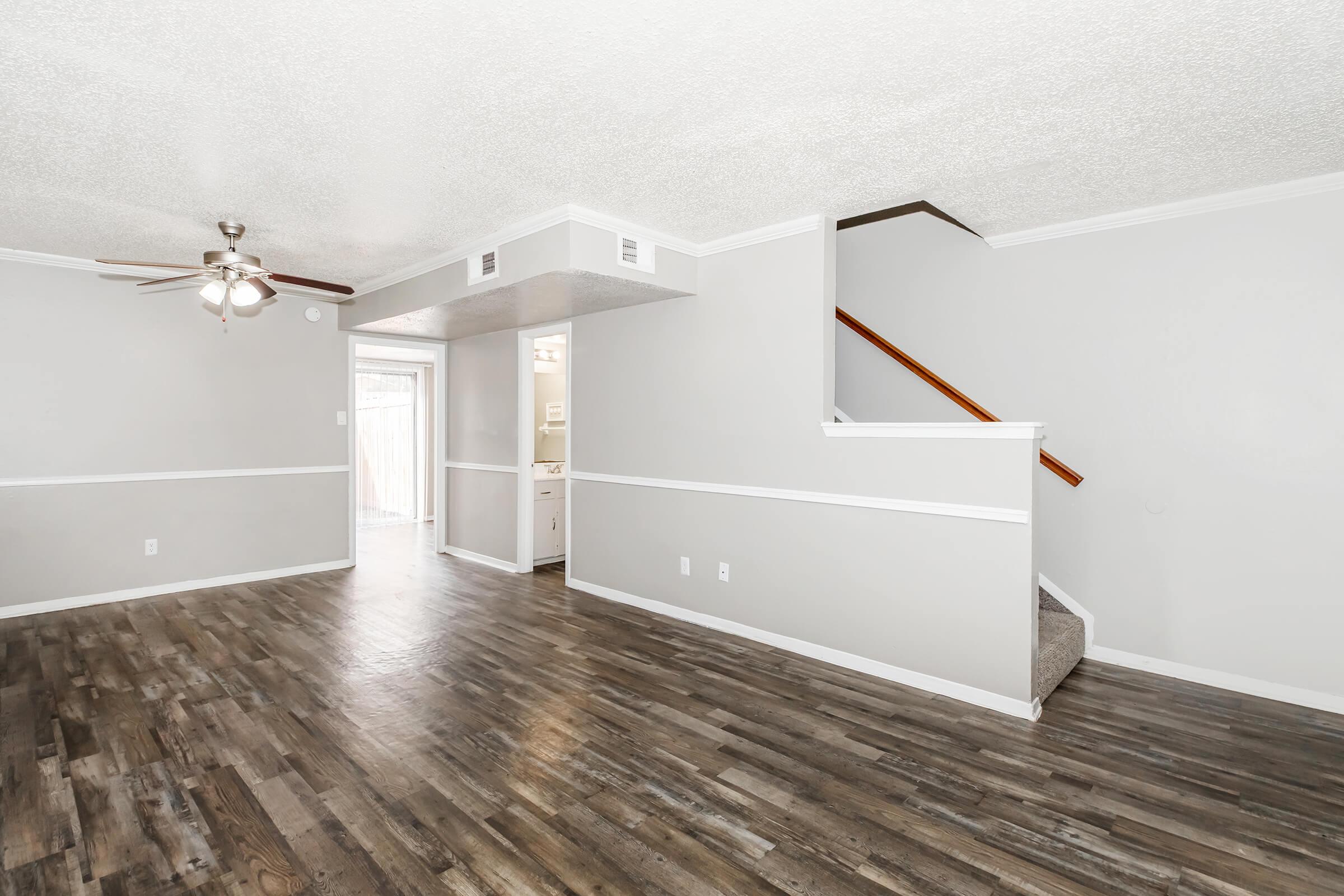 Spacious living room with neutral gray walls and wood-like flooring. A ceiling fan is mounted above, and a staircase leads to an upper level. To the left, there is an entryway that opens to a kitchen area. Natural light comes from a sliding glass door at the back. White wainscoting adds a decorative touch.