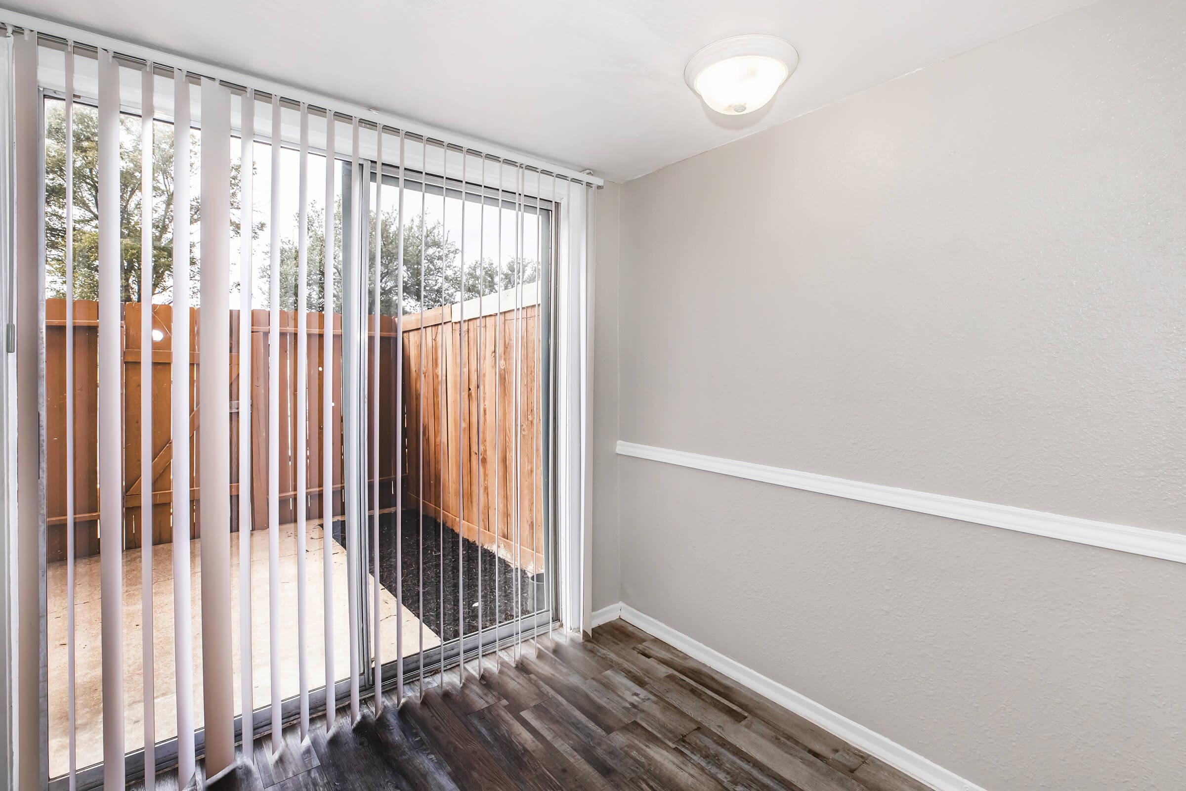 A well-lit room featuring a sliding glass door with vertical blinds leading to an outdoor area. The walls are painted in a light gray, and there is a decorative white trim. The floor has a wood-like finish, and a wooden fence is visible outside through the door.