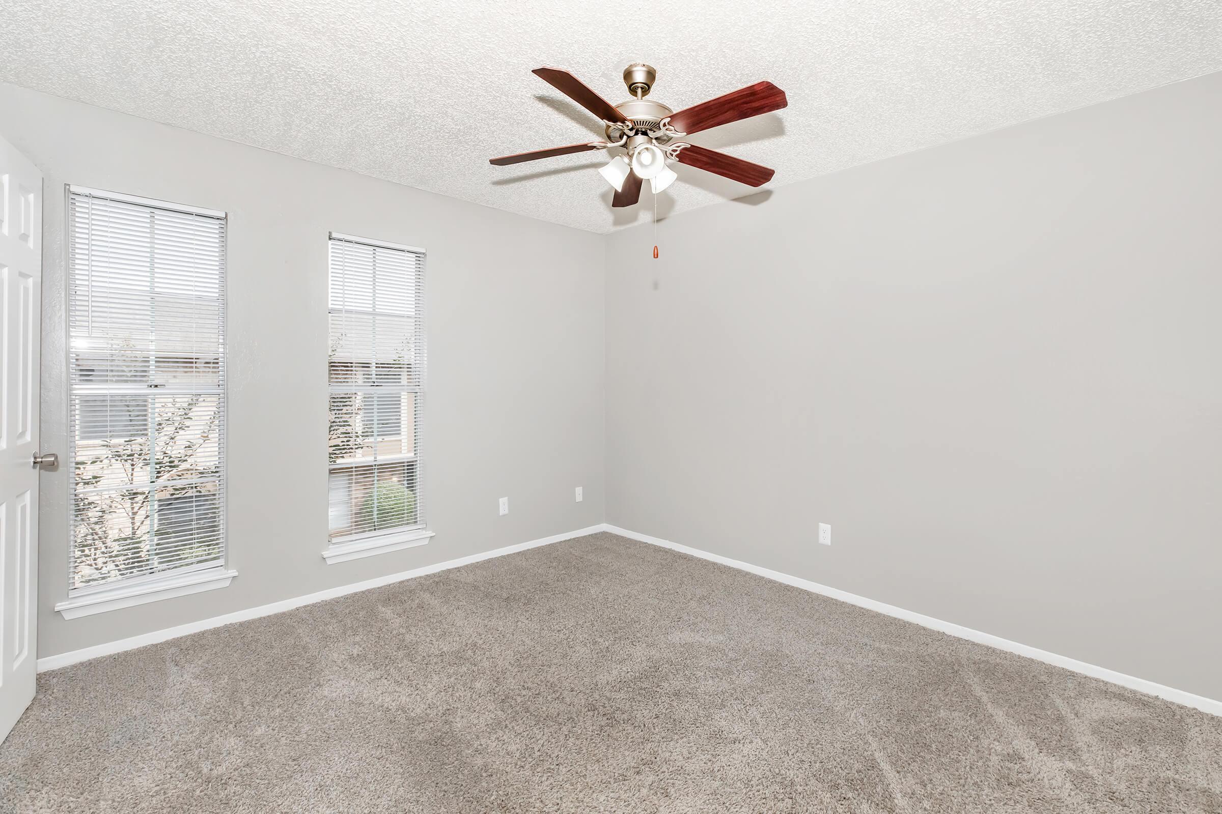 A clean, empty room featuring a ceiling fan with wooden blades, two windows allowing natural light, and light grey walls. The floor is carpeted in a neutral tone, creating a spacious and inviting atmosphere.