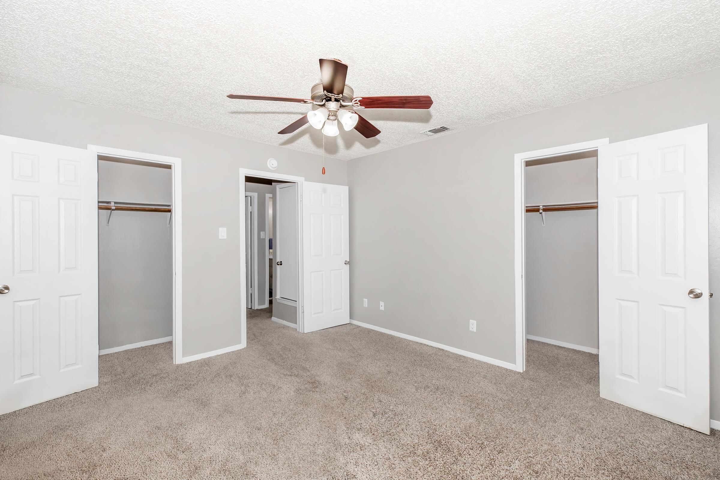 A spacious, empty bedroom with light gray walls and a textured ceiling. The room features a ceiling fan with wooden blades, two open closet doors on the sides, and a doorway leading to another room in the background. The carpet is beige and plush, contributing to a cozy atmosphere.