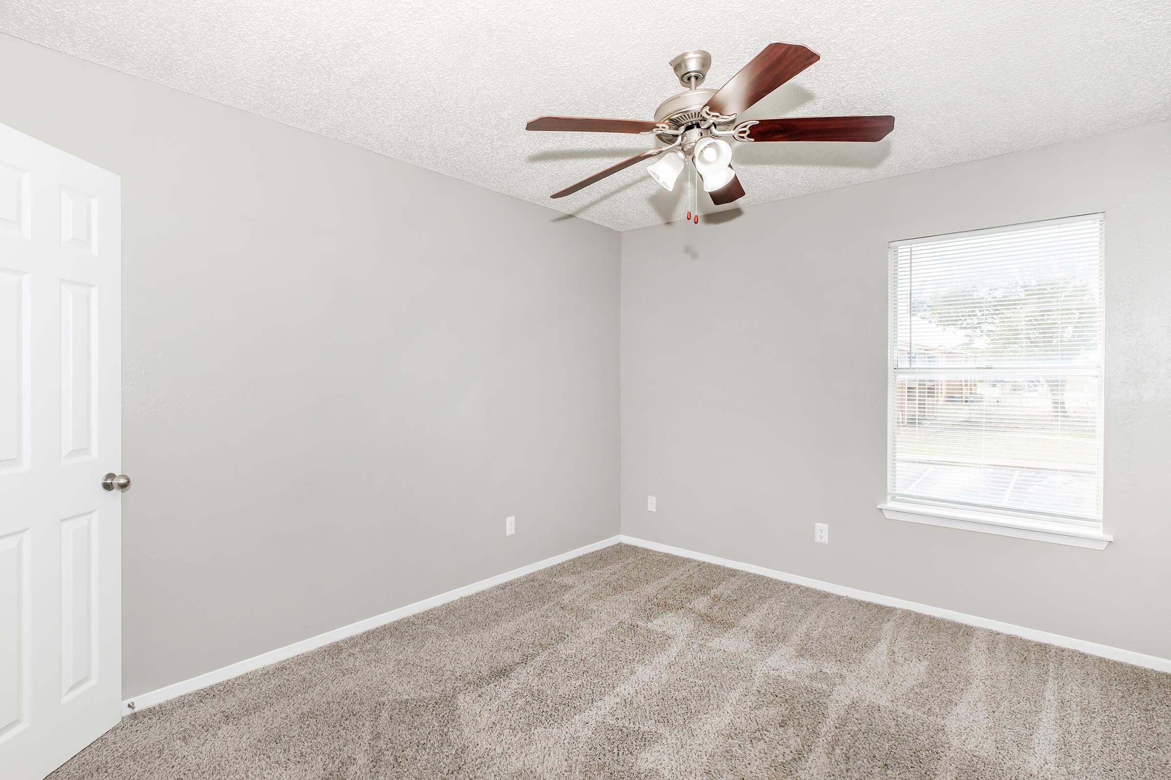 A spacious, empty room featuring light-colored walls and carpet. The room has a window allowing natural light, and a door on the left. The ceiling is slightly textured, and there are no furniture items present, creating a clean, minimalist atmosphere suitable for various uses.
