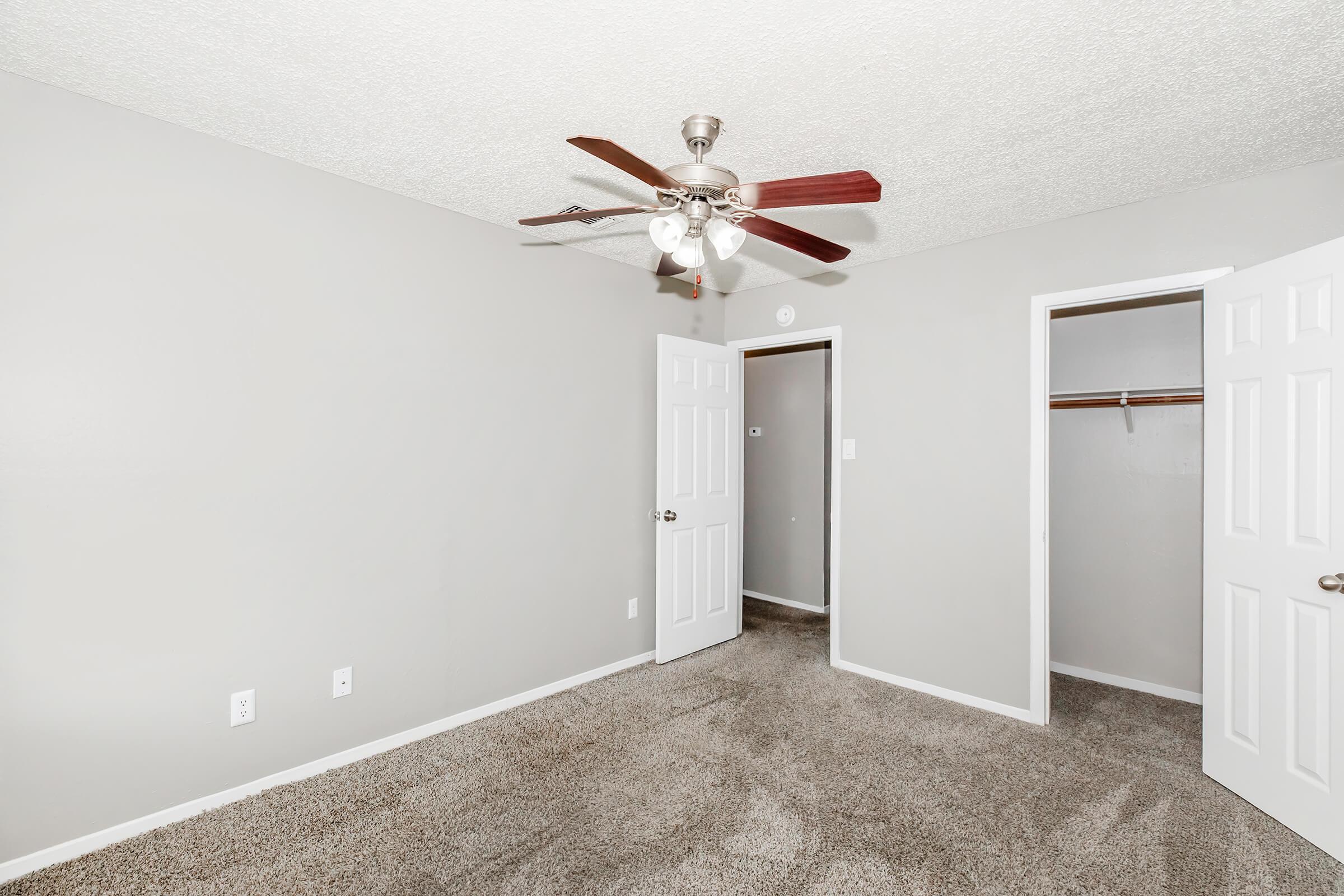 A spacious room with gray walls and carpeted flooring. It features a ceiling fan with light, two white doors—one leading to a closet and the other to another room. The room is well-lit and empty, offering potential for various uses.