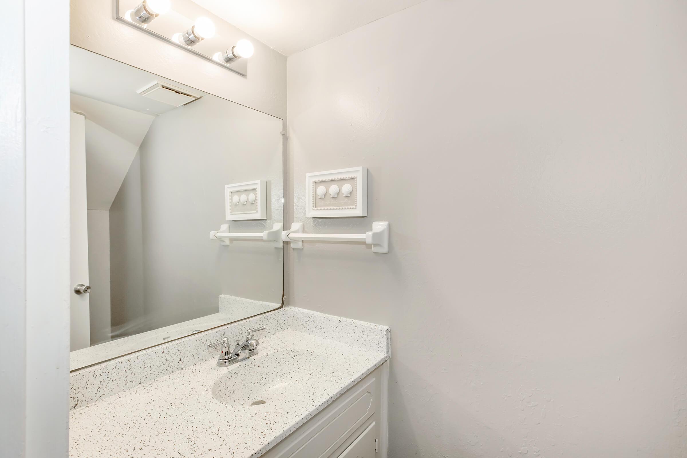A modern bathroom with a white sink and countertop, a large mirror above the sink, and two white towel racks on the wall. The walls are painted in a light neutral color, giving the space a clean and simple appearance. Bright lighting is provided by three bulbs above the mirror.