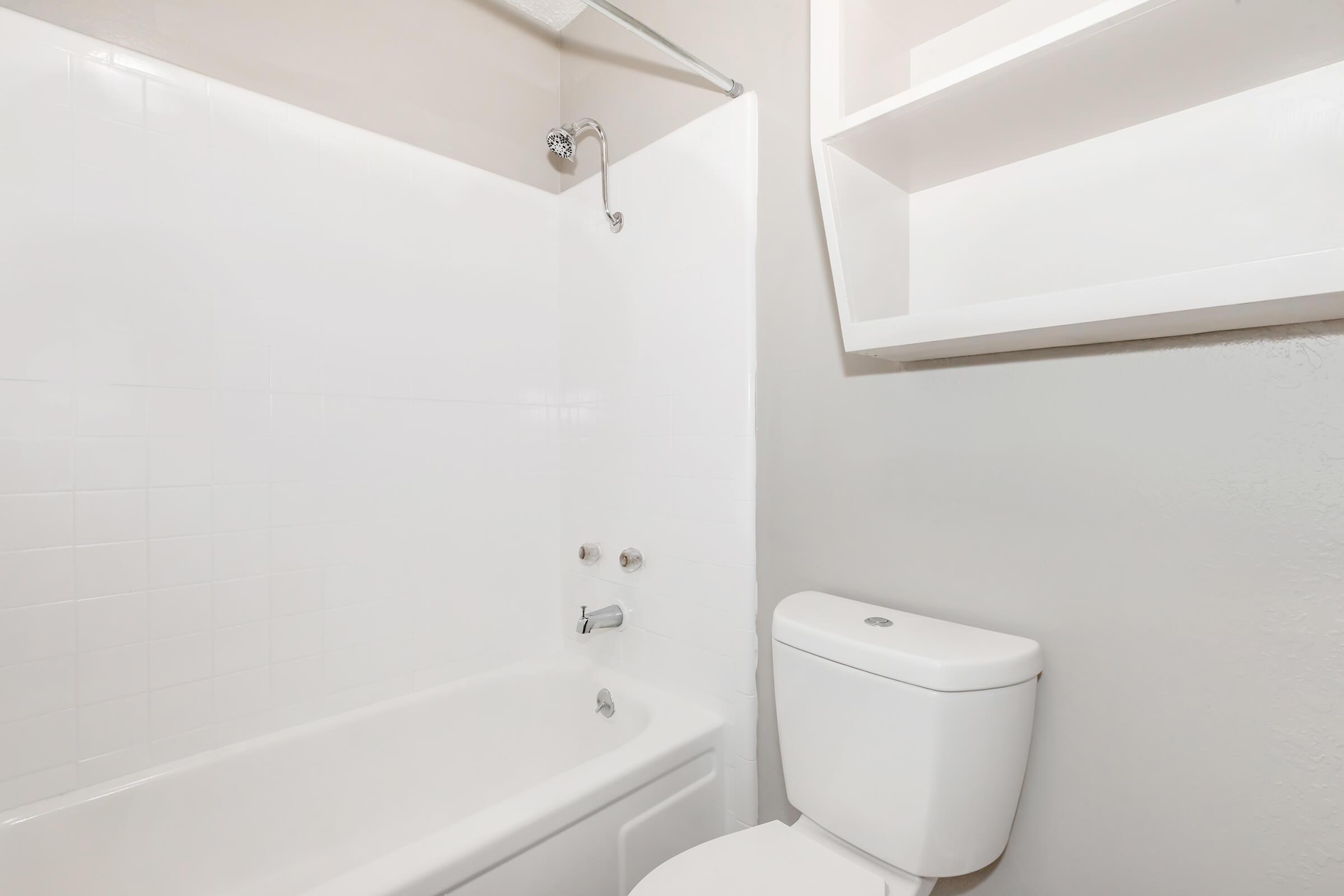 A simple bathroom featuring a white bathtub with a showerhead, a white toilet, and a small shelf above the bathtub. The walls are a light gray color, and the overall space appears clean and modern.