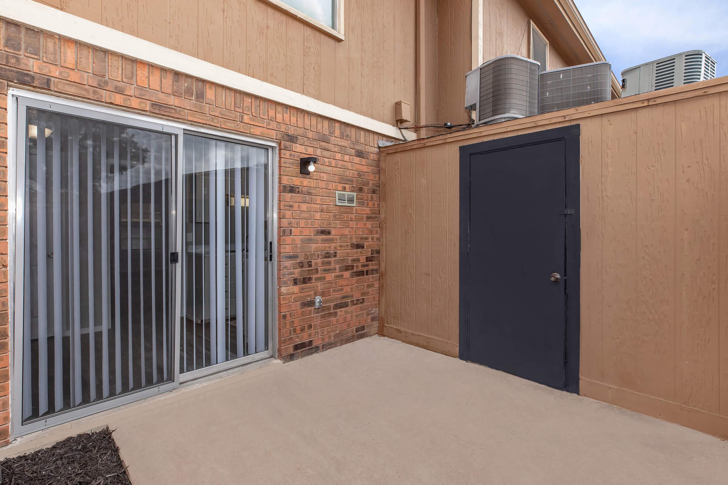 Exterior view of a house featuring a small patio area with a sliding glass door on one side, a solid dark door on the other, and brick walls. An air conditioning unit is mounted on the exterior. The ground is concrete, and there is a small patch of dark mulch nearby.