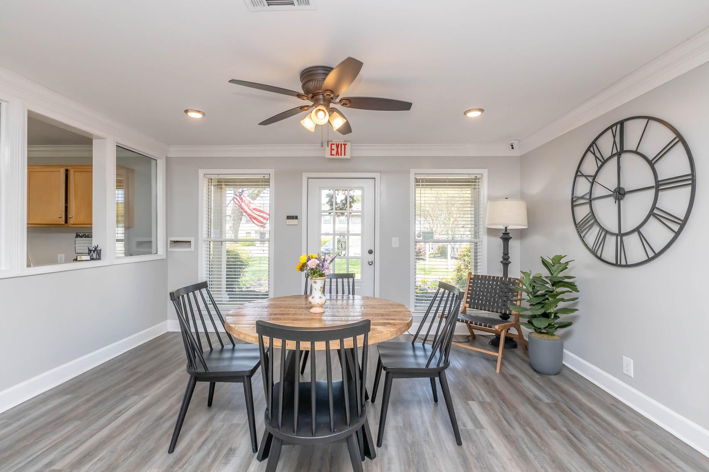 A modern dining area featuring a round wooden table with black chairs, a ceiling fan, and a large wall clock. Bright natural light enters through windows with blinds, and there are decorative plants and a vase of flowers on the table. An American flag is visible outside.