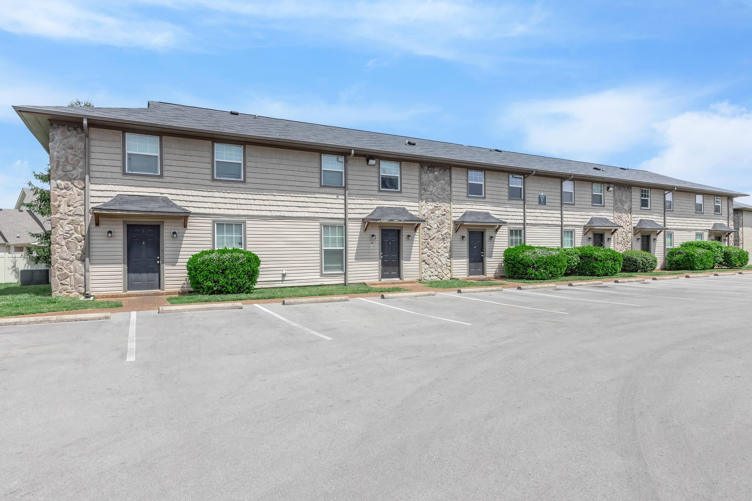 Row of beige multi-family apartments with stone accents, featuring black doors and windows. Installed greenery is visible in front of the buildings. A paved parking lot is present in the foreground, with empty parking spaces. The sky is clear and blue, with a few scattered clouds.