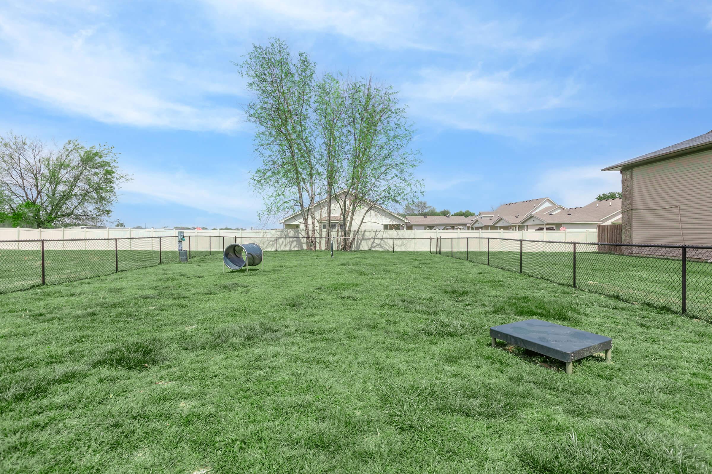 A grassy yard enclosed by a black fence, featuring a small dog house on the left side and a raised platform in the center. There are a few trees in the background and houses visible on the right side under a blue sky.
