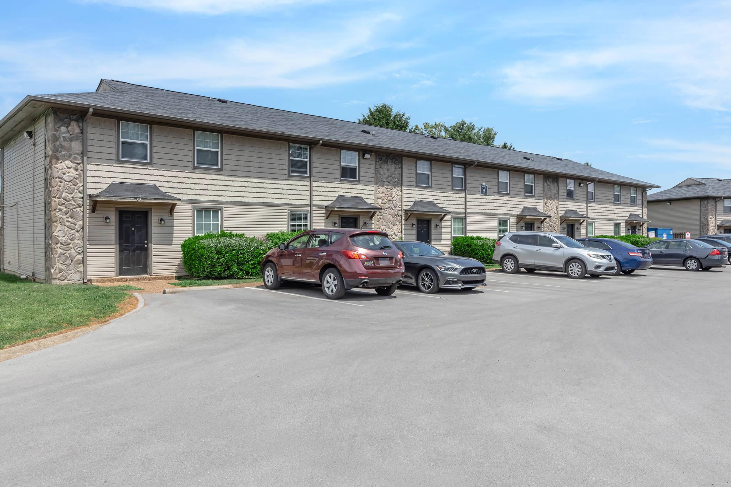 Row of multi-unit residential buildings with a mix of cars parked in front. The buildings feature a light-colored exterior with stone accents. Green bushes line the façade, and a clear blue sky is visible above. The scene suggests a well-maintained suburban neighborhood.
