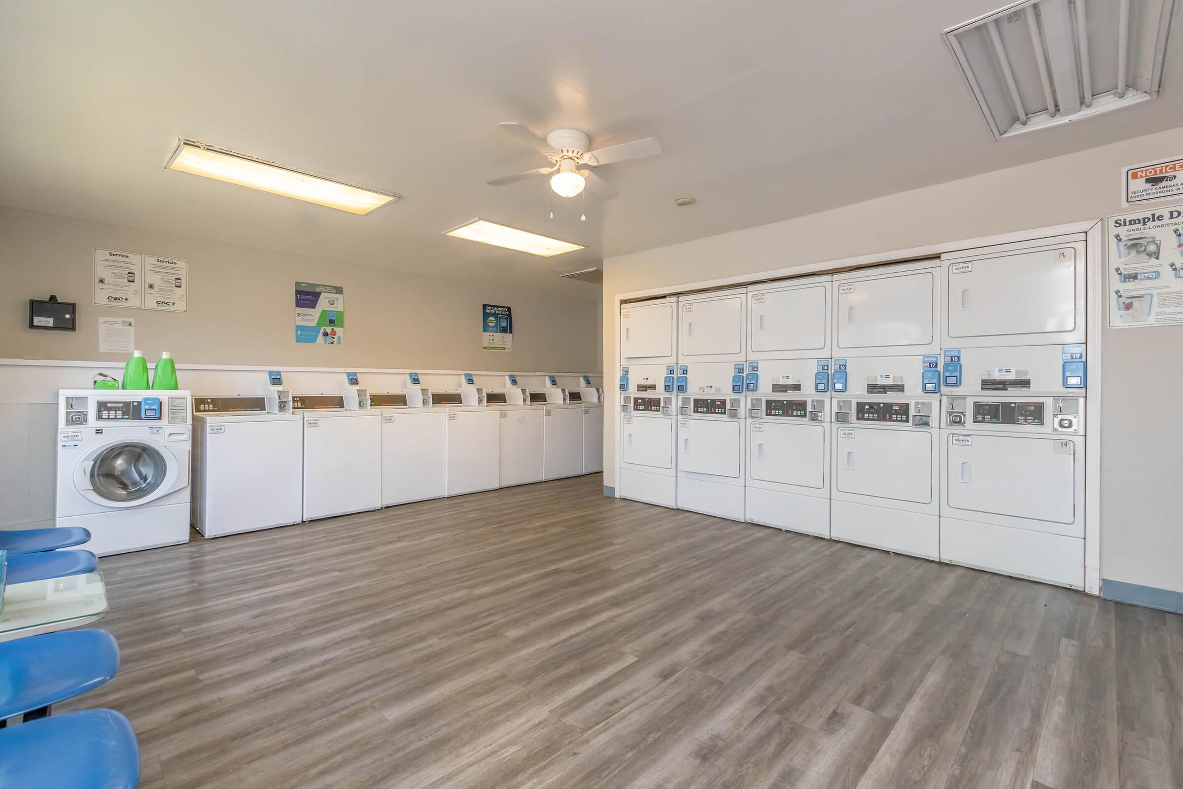 A clean, well-lit laundry room featuring multiple white washing machines and dryers arranged in rows. There are blue folding chairs along one wall, and informational posters are displayed. A ceiling fan provides air circulation in the modern space.