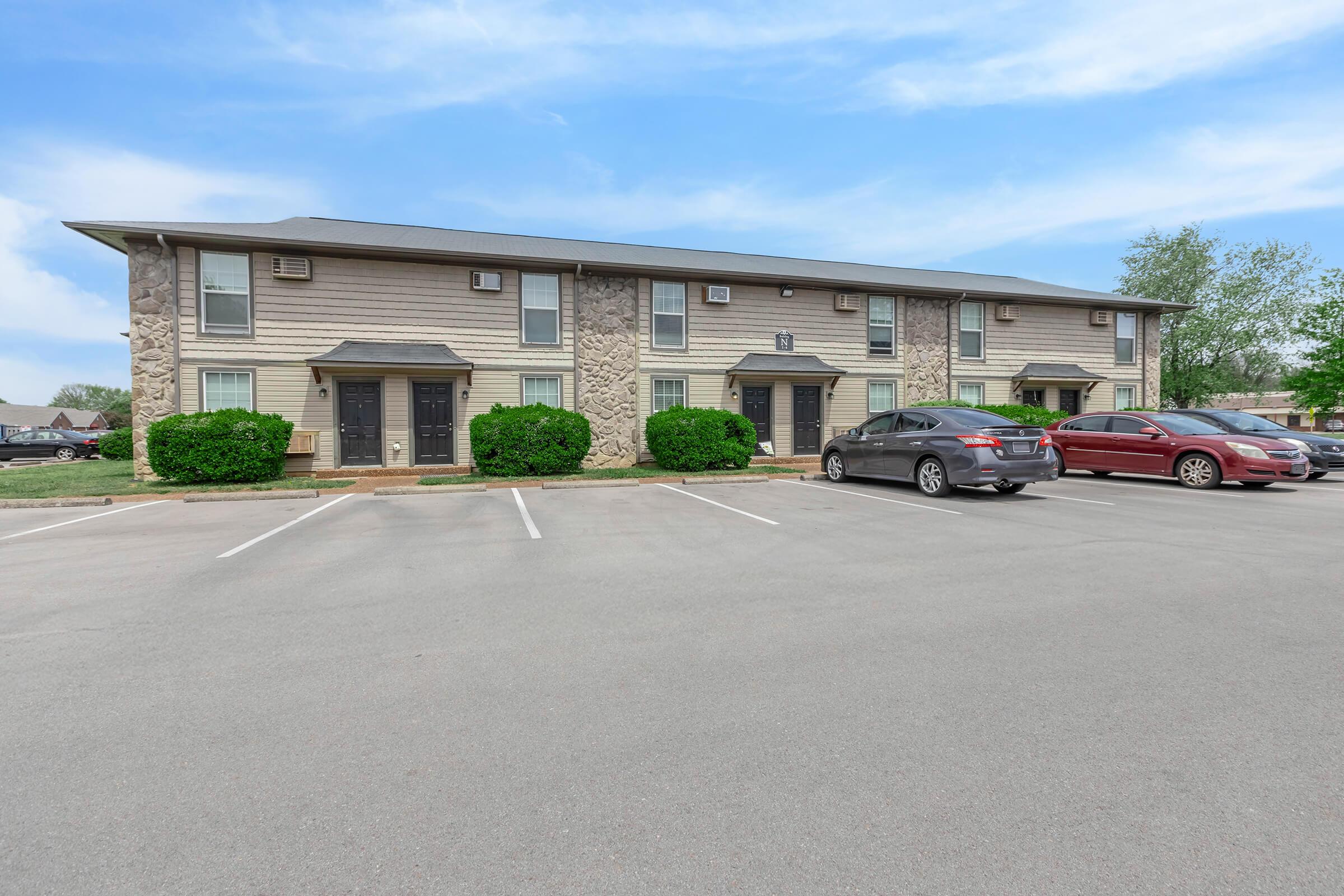 A two-story apartment building with stone accents, featuring multiple entrances. Two cars are parked in front, one gray and one red. The area is well-maintained with green shrubs and a clear blue sky overhead. Parking spaces are visible in front of the building.