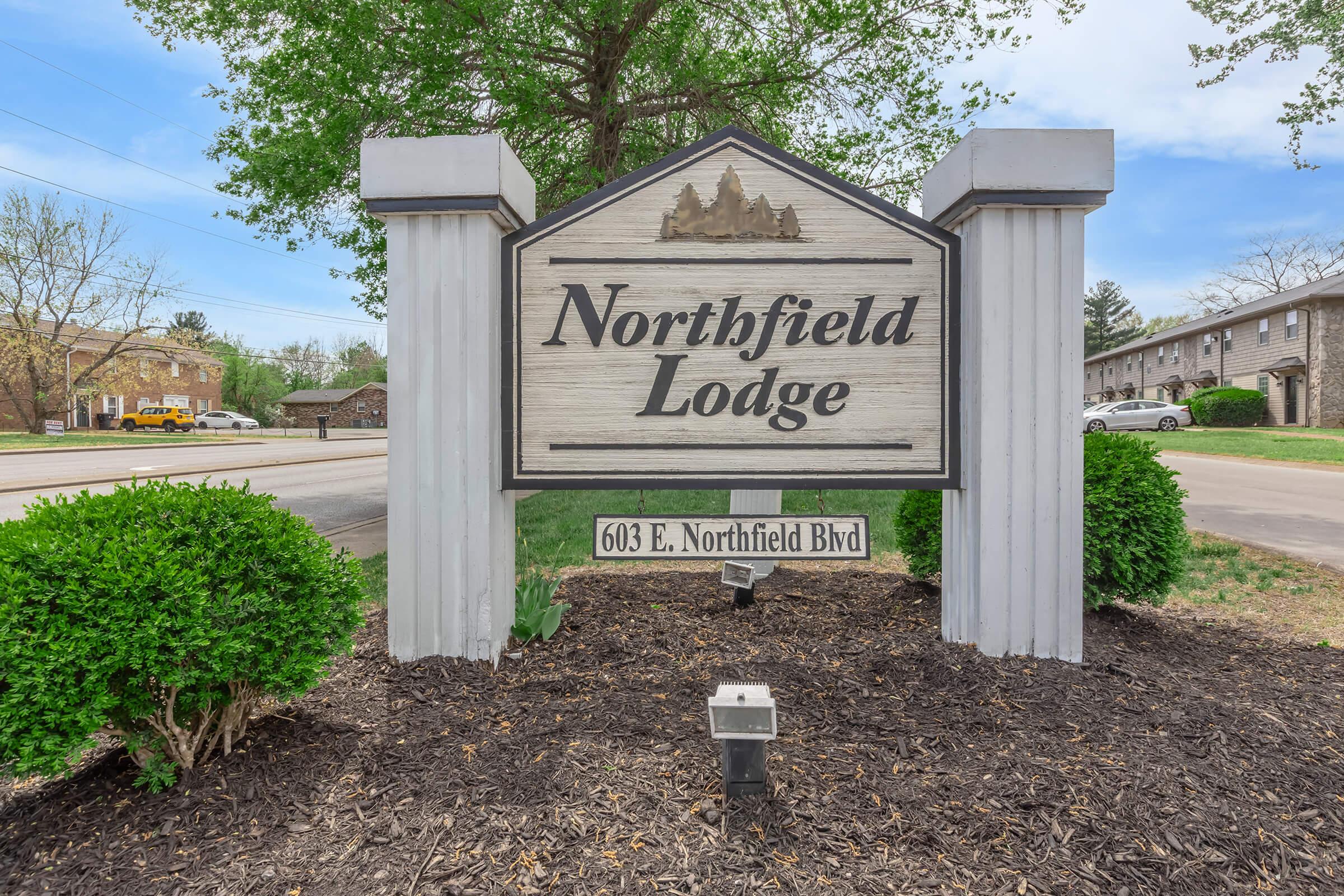 Sign for Northfield Lodge, featuring a wooden design with "Northfield Lodge" inscribed at the top and the address "603 E. Northfield Blvd" below. Surrounding the sign are neatly trimmed bushes, and the background shows a street and trees under a slightly cloudy sky.