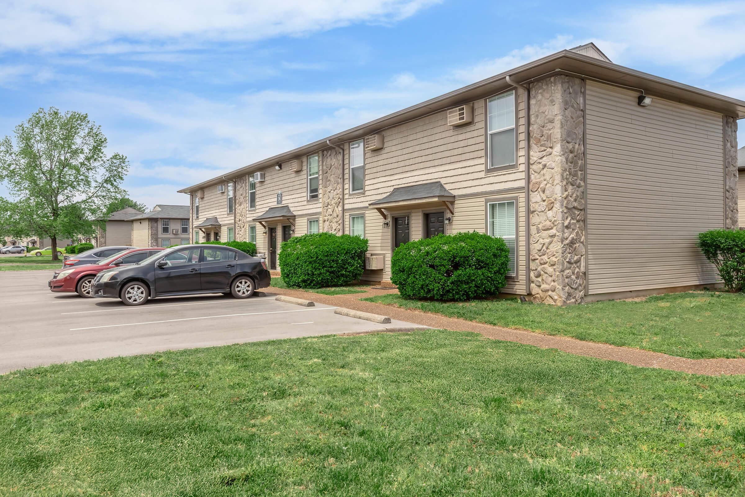 Exterior view of a residential apartment complex featuring two-story buildings with stone accents. A grassy area with neatly trimmed bushes surrounds the buildings, and a parking lot with several parked cars is visible in front. The sky is clear with few clouds.