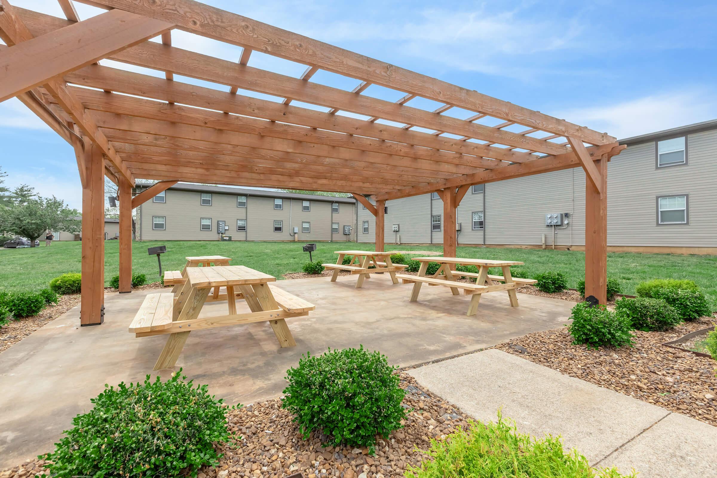 An outdoor gathering area featuring a wooden pergola with picnic tables and grill stations, surrounded by well-manicured shrubs and grass, under a clear blue sky.