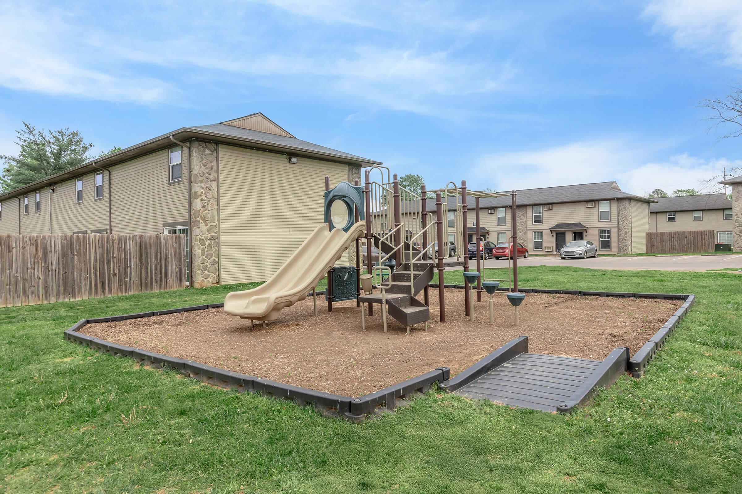 A playground with a slide, climbing structure, and various play equipment surrounded by a grassy area, with a wooden fence in the background and residential buildings nearby. The sky is clear with a few clouds.
