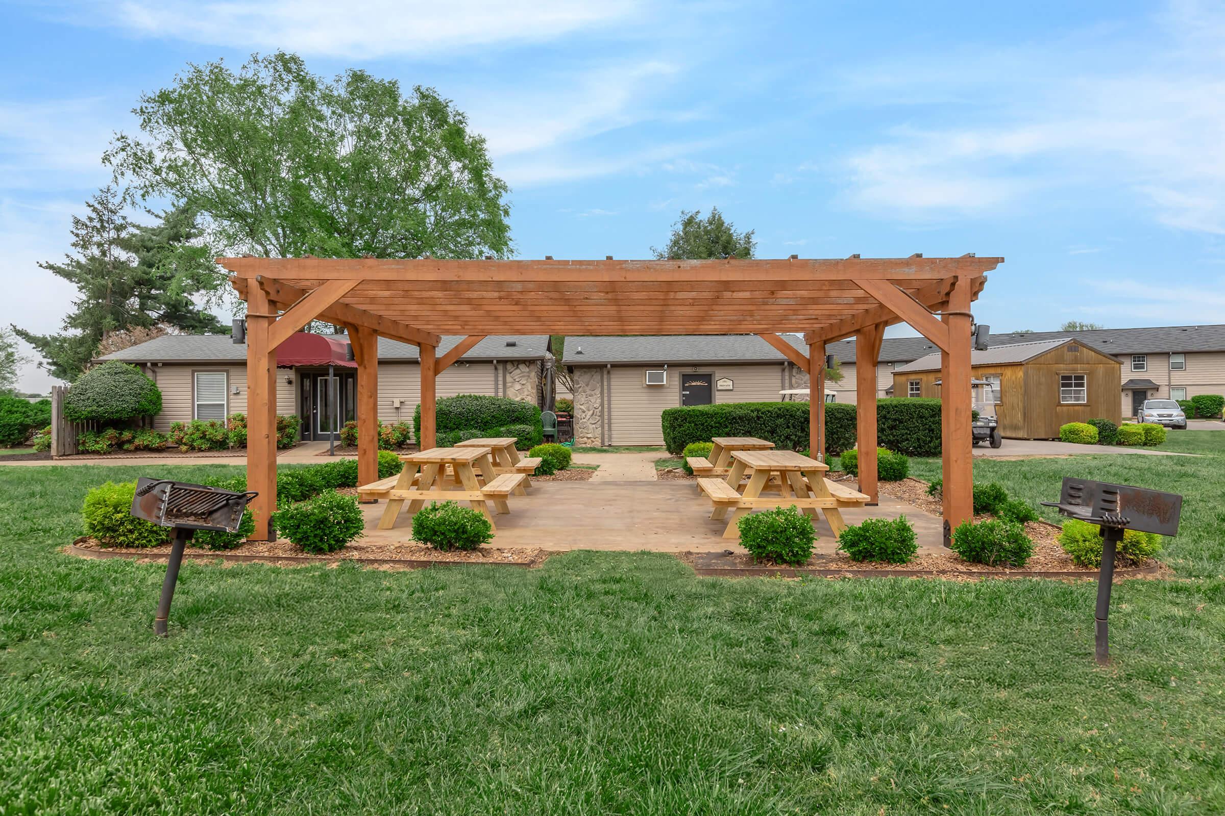 A wooden pergola with picnic tables underneath, surrounded by manicured grassy areas and shrubs. There are barbecue grills on either side. In the background, there are residential buildings with a clear blue sky above.