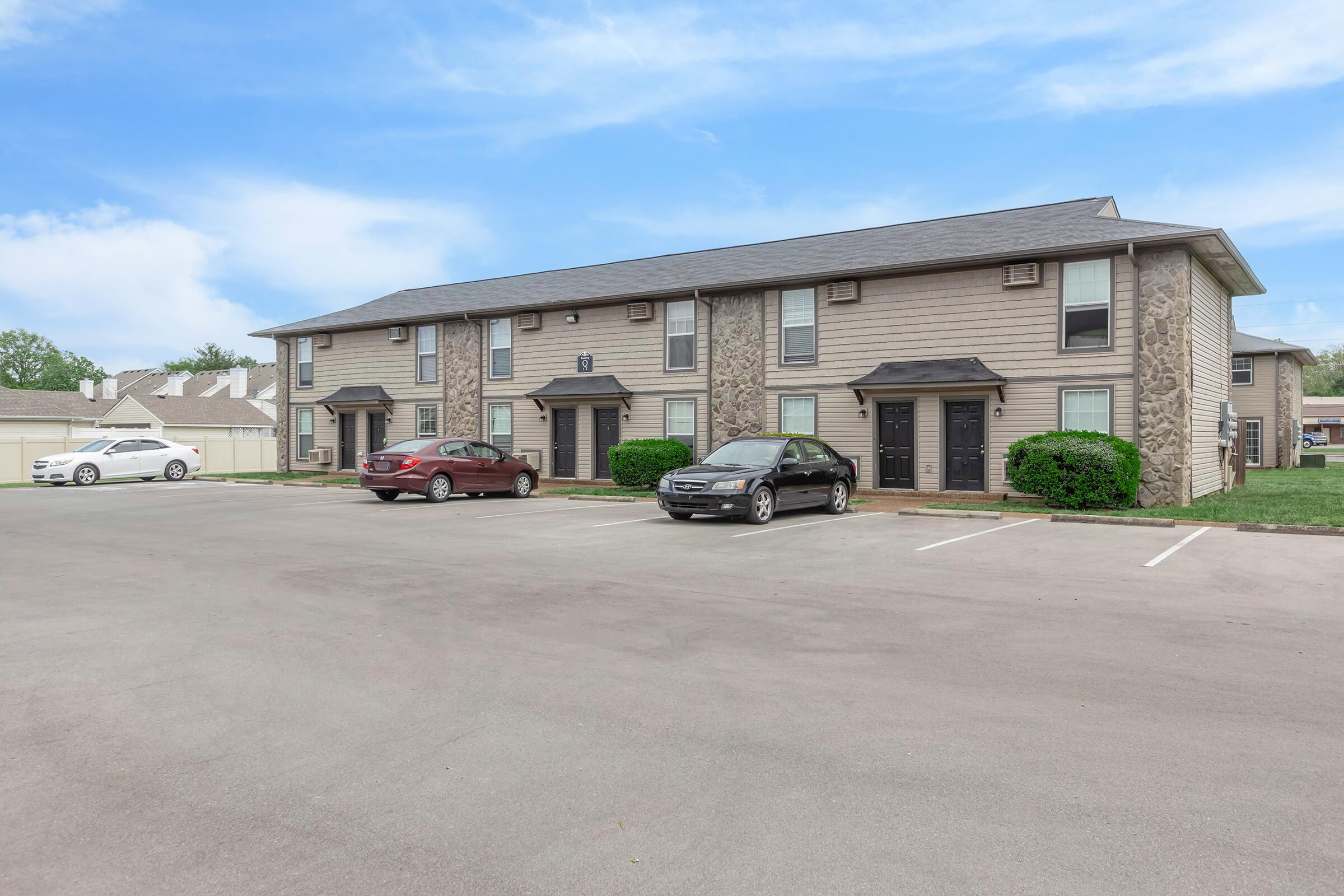 Row of multi-unit residential buildings with a light grey exterior, featuring stone accents. Several parked cars, including a white vehicle and two shades of grey, are in the foreground. Well-kept lawns and a clear blue sky create a welcoming atmosphere.