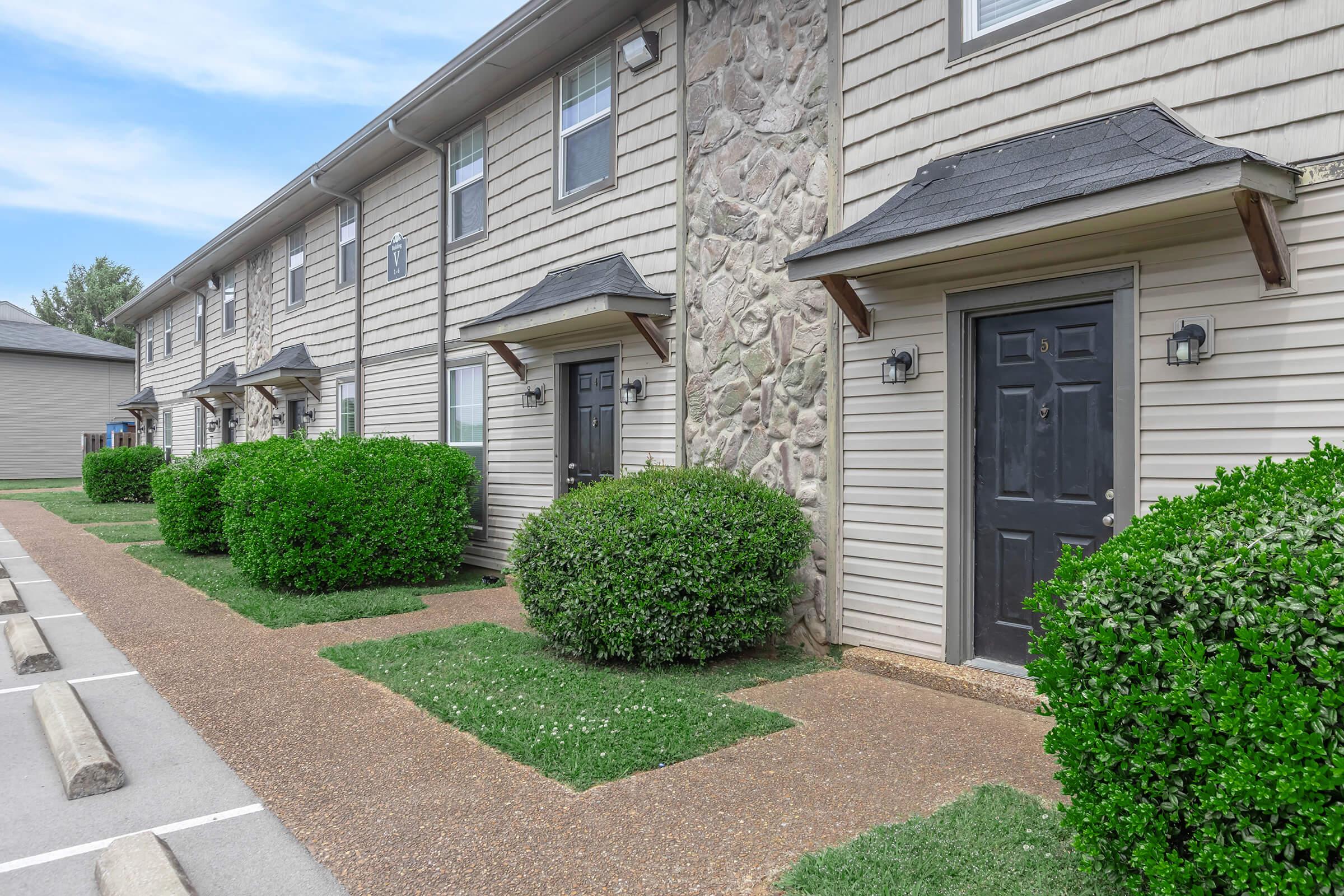 A row of apartment buildings featuring multiple units with gray siding and stone accents. The entrance doors are black and have stylish light fixtures. Neatly trimmed bushes line the pathway, and there is a gravel walkway in front of the units, with parking spaces visible in the foreground.