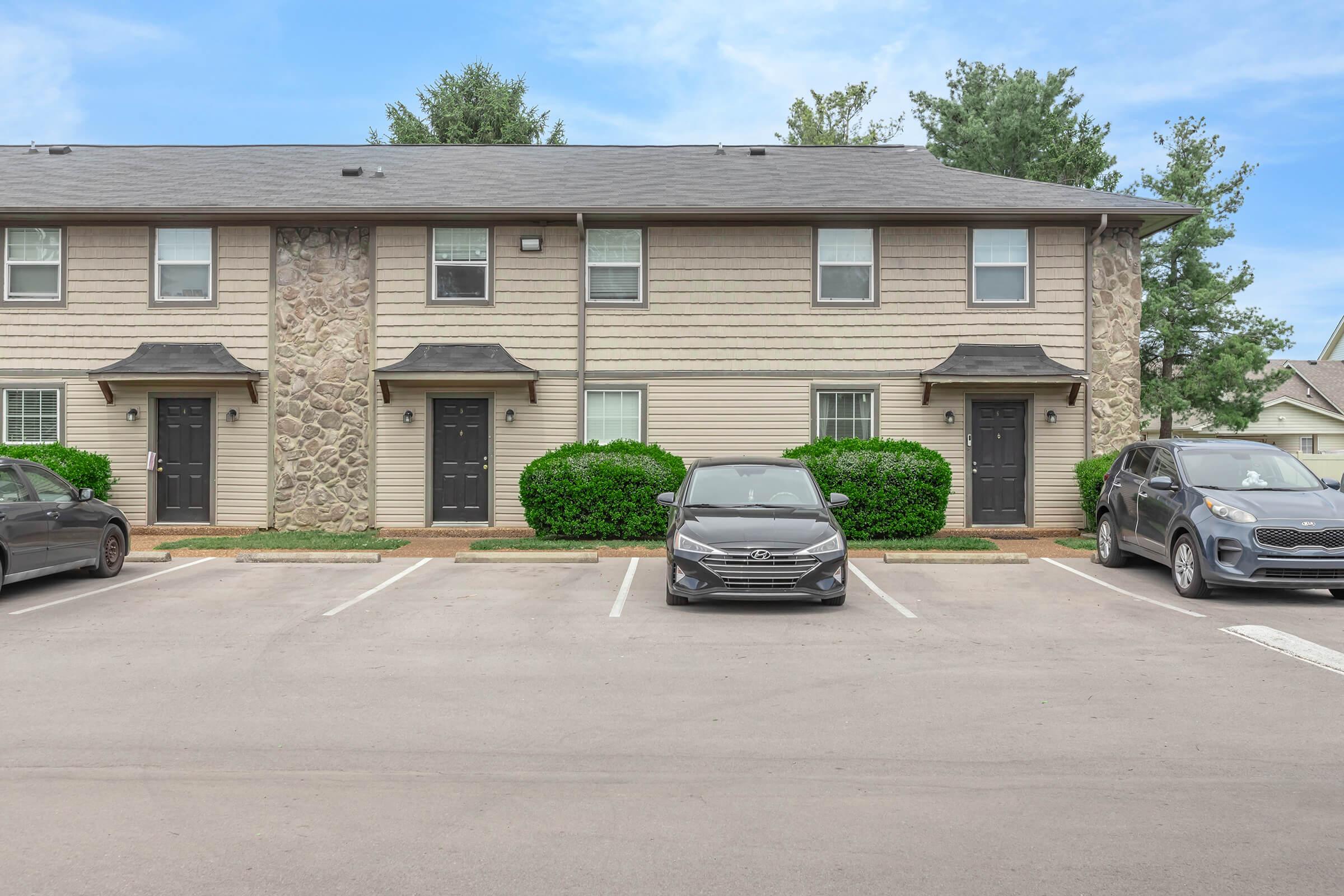 Two-story residential building with a beige exterior featuring stone elements. There are four doors, two on each floor. The parking area in front has three parked cars, with greenery surrounding the entrances. The sky is clear and the scene appears well-maintained.