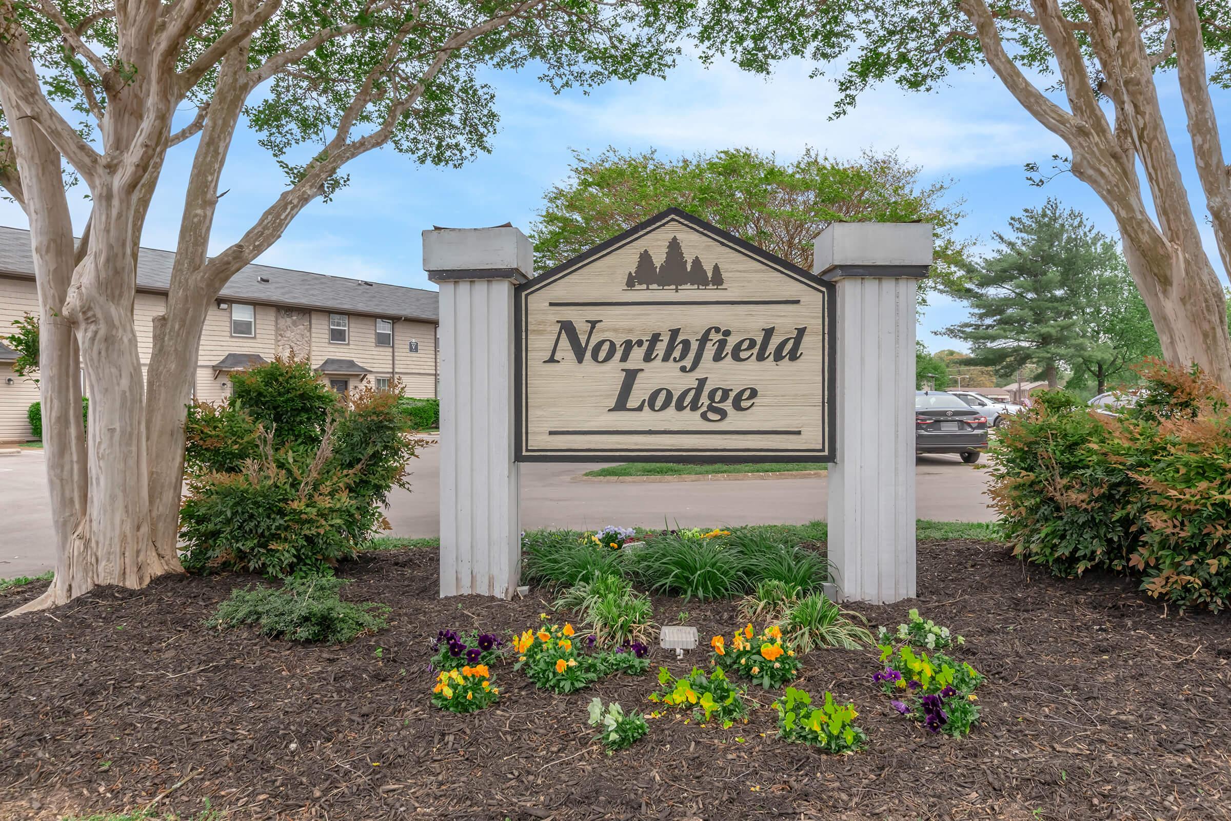 Sign for Northfield Lodge surrounded by colorful flower beds and green shrubs, with trees in the background. The lodge appears to be part of a residential area, with parked cars visible nearby. The sky is clear and blue, indicating a pleasant day.