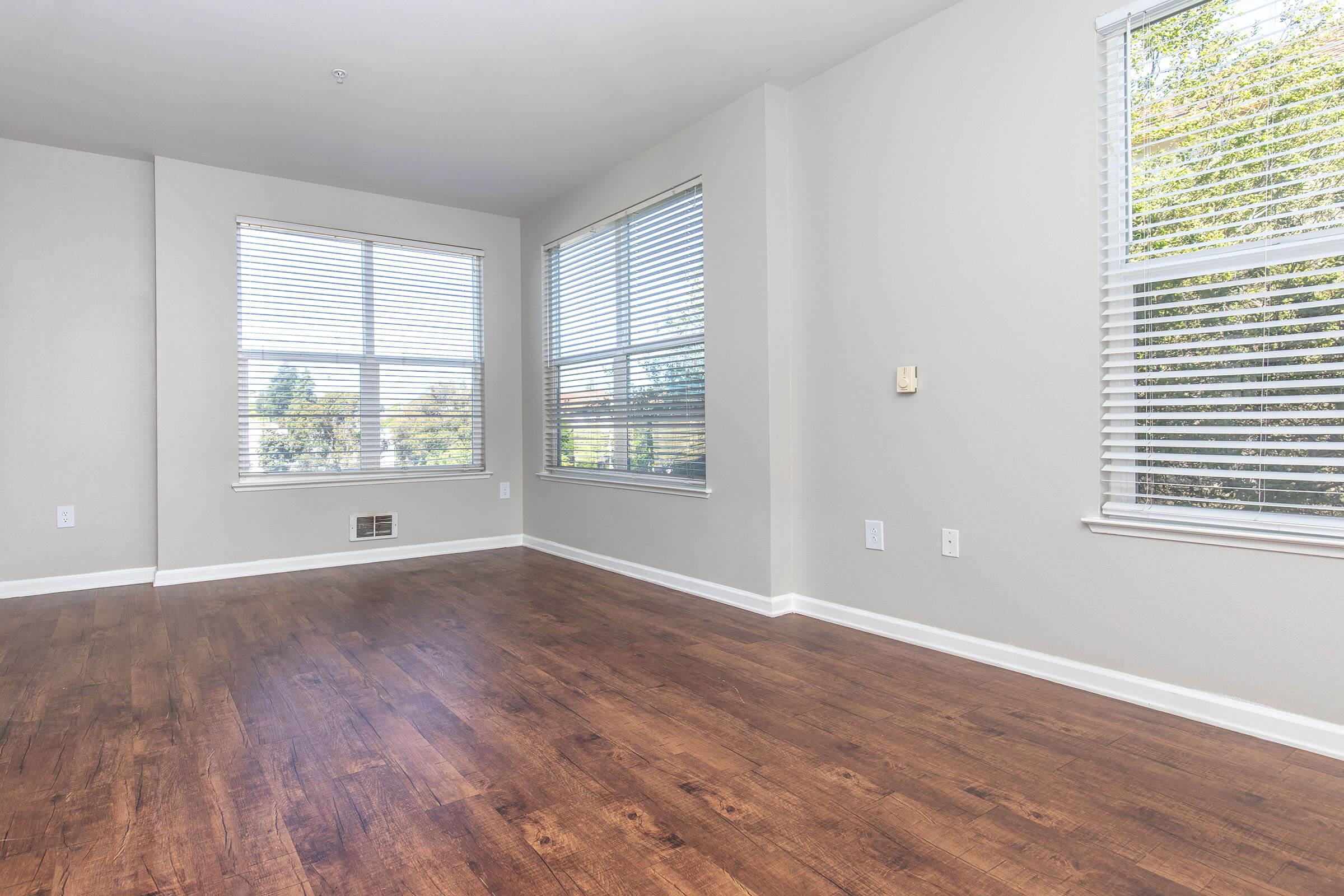 A bright, empty room with large windows featuring white blinds. The walls are painted light gray, and the floor is a rich, polished wood. Natural light floods the space, highlighting the clean lines and modern feel.