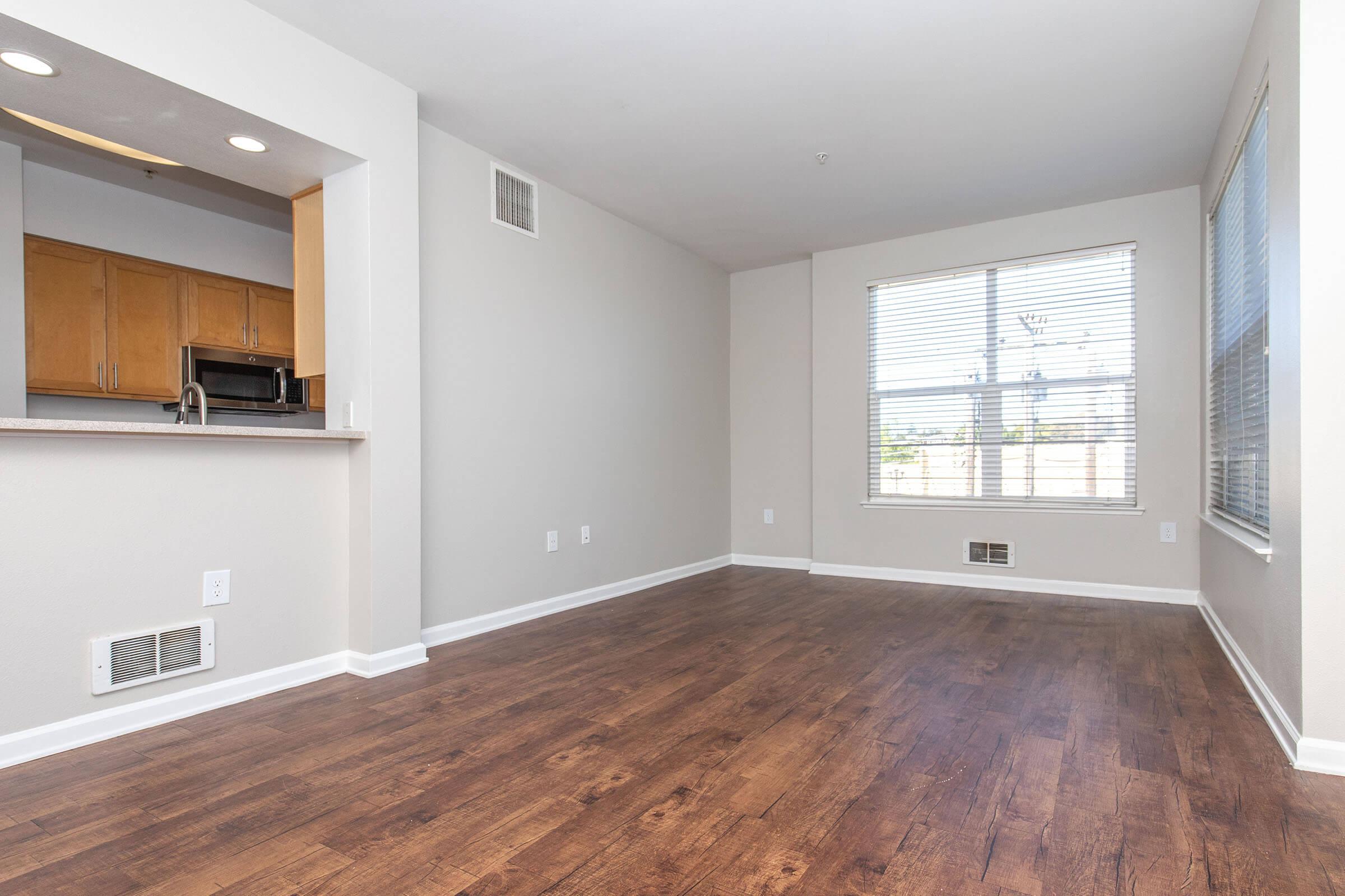 A spacious, empty living room with light-colored walls and wooden flooring. The room features a large window with blinds allowing natural light, and a doorway leading to a kitchen area with wooden cabinets visible in the background. No furniture is present, emphasizing the room's openness.